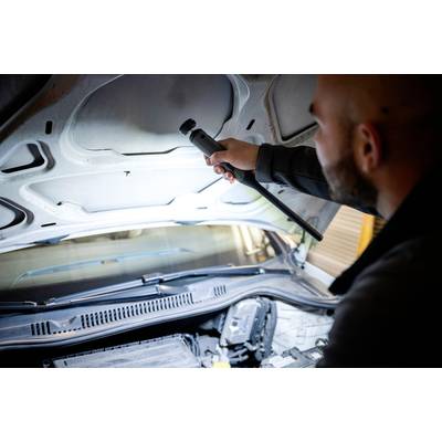 Ein Mensch untersucht den Motorraum eines Autos mit einer Taschenlampe, möglicherweise bei einer Wartung oder Inspektion.