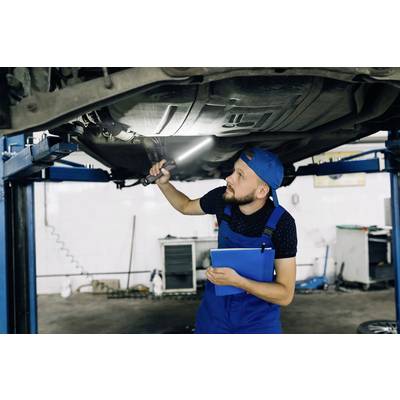 Ein Mechaniker in blauer Arbeitskleidung untersucht mit einer Lampe das Unterteil eines Autos, das auf einer Hebebühne steht.