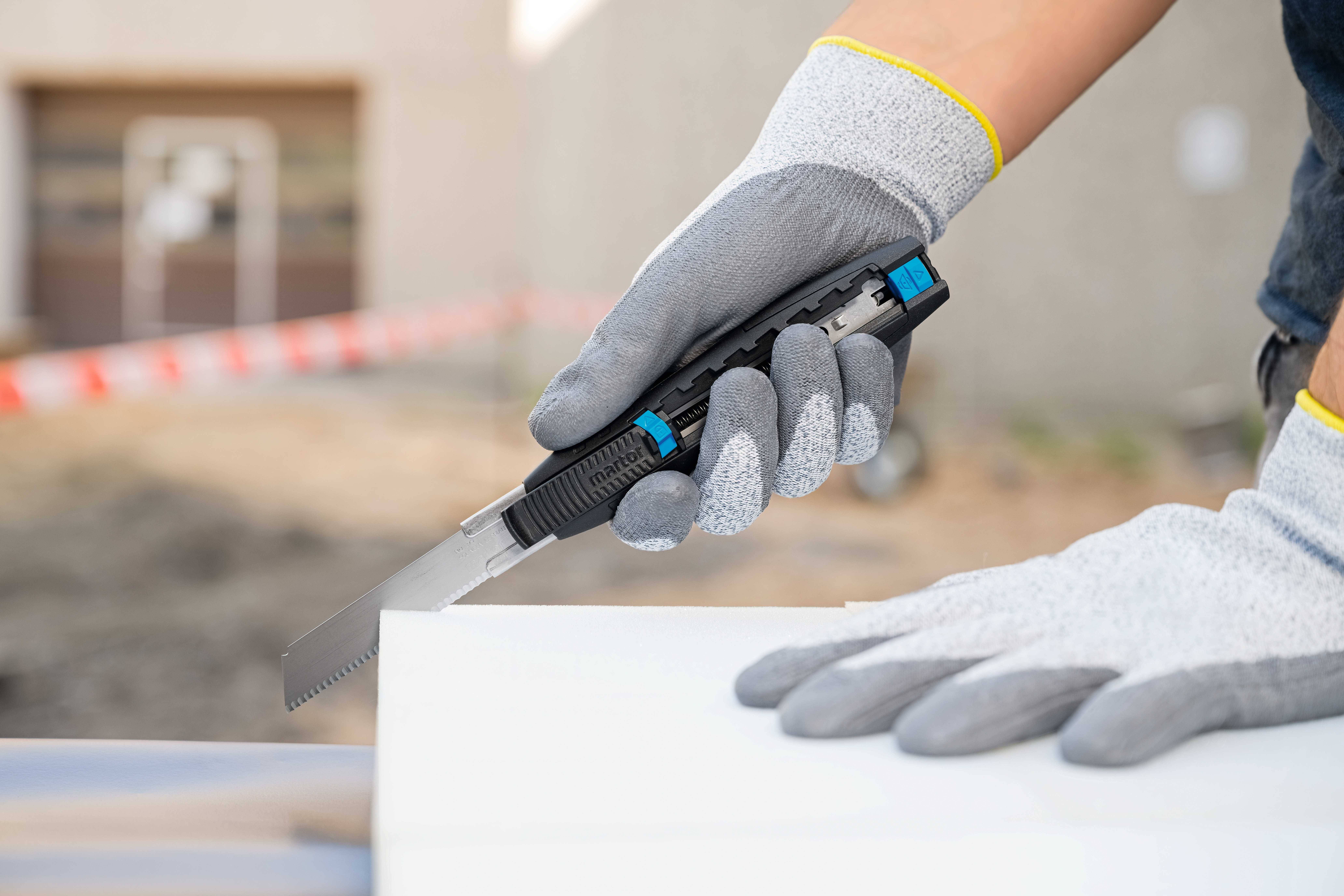 Eine Person mit grauen Handschuhen benutzt einen Teppichmesser, um ein weißes Material zu schneiden. Hintergrund unscharf.