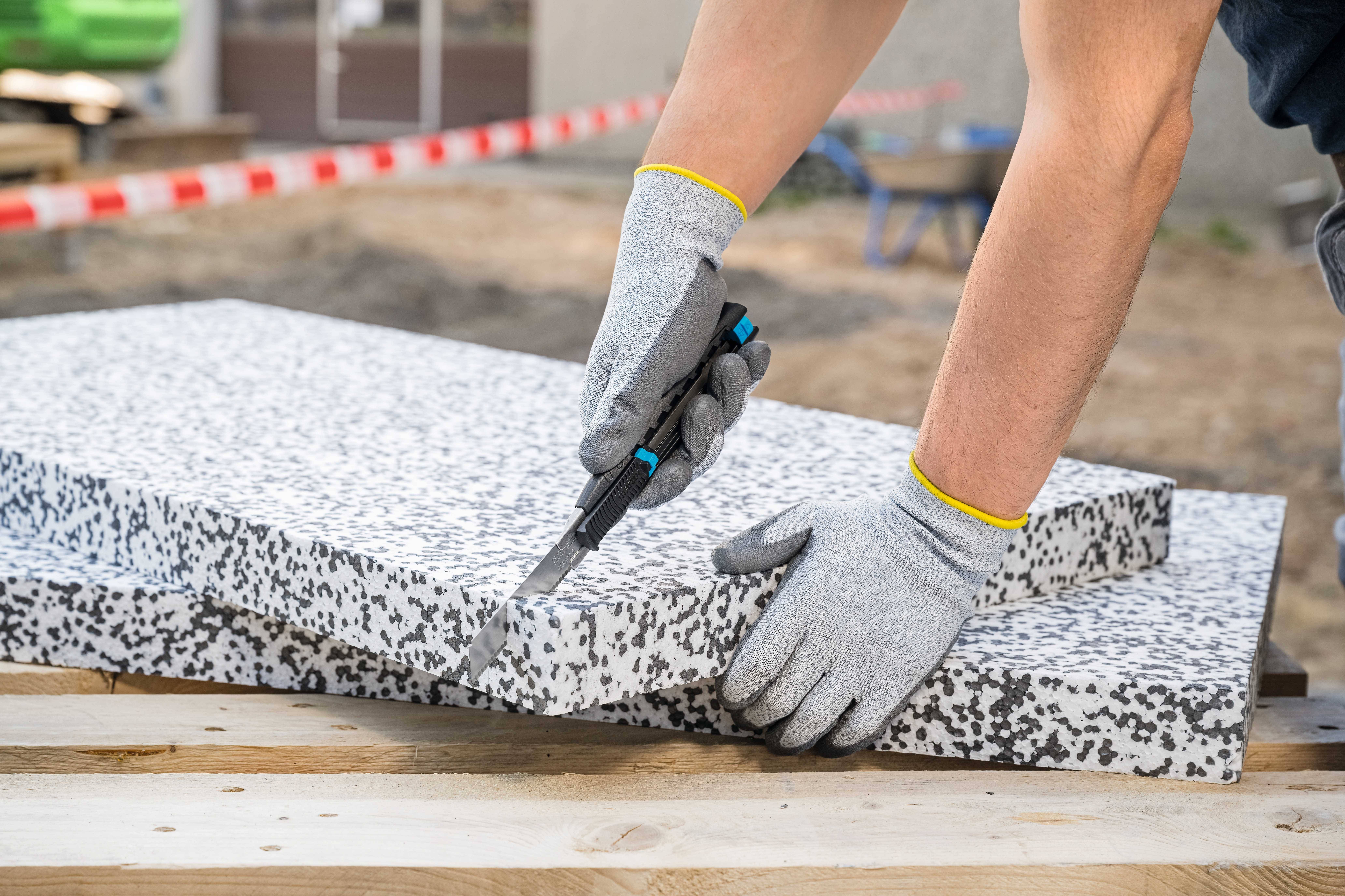 Eine Person mit grauen Handschuhen bearbeitet mit einem Werkzeug die Kante einer grob gepunkteten Steinplatte auf einer Baustelle.