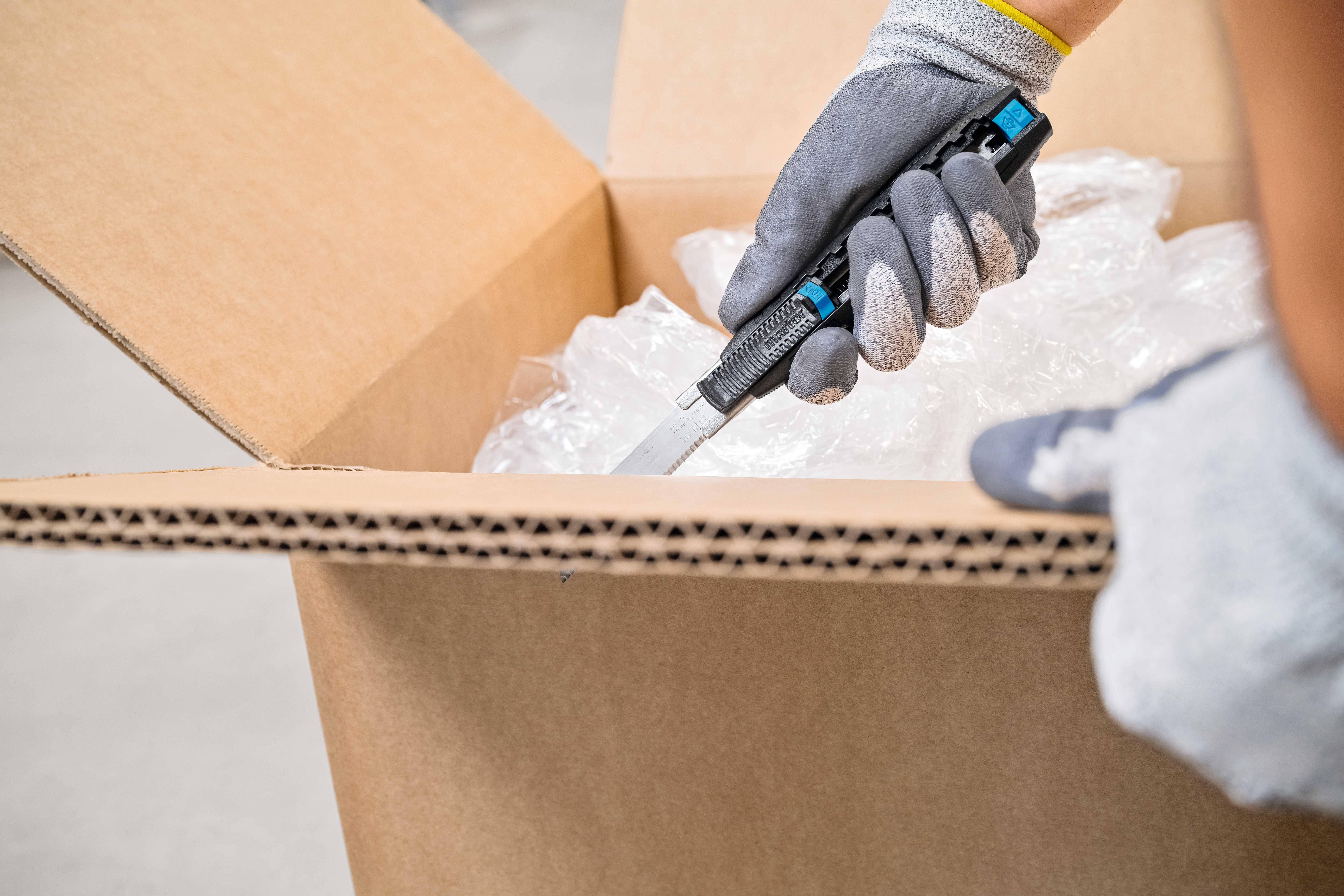 Eine Person mit grauen Handschuhen öffnet vorsichtig einen Pappkarton mit einem Cuttermesser. Der Karton enthält Luftpolsterfolie.