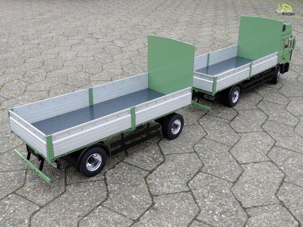 Ein grüner Lastwagen mit Anhänger, beide offen und leer, steht auf einer gepflasterten Fläche. Sie haben hohe Seitenwände und sind bereit für den Transport.
