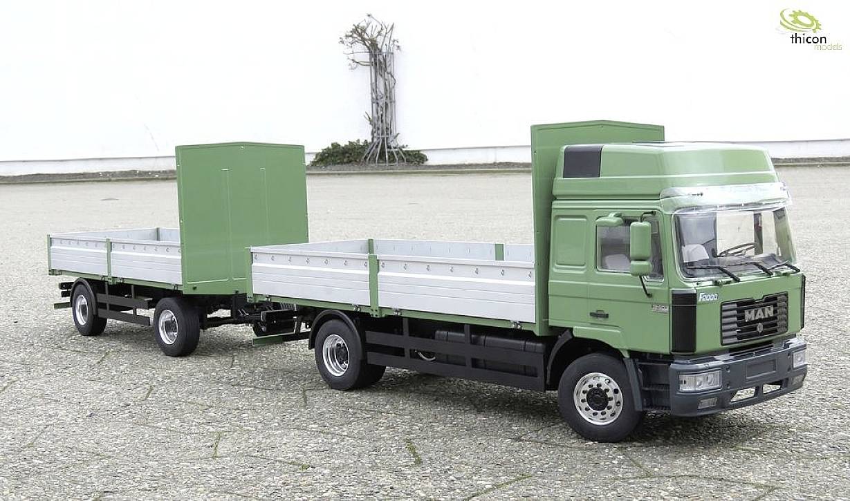 Grüner LKW mit Anhänger steht auf gepflastertem Platz, im Hintergrund weiße Wand mit Kletterpflanze; keine Personen zu sehen.