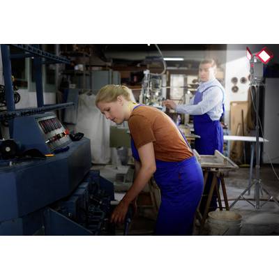 Eine Frau und ein Mann arbeiten in einer Werkstatt an Maschinen. Beide tragen blaue Overalls. Im Hintergrund sind Werkzeuge zu sehen.