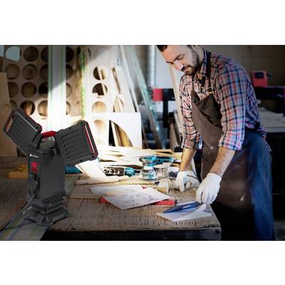 Ein Mann arbeitet in einer Werkstatt und nutzt Werkzeuge auf einem Tisch. Im Vordergrund steht eine Lampe, die den Bereich beleuchtet.