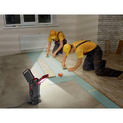Zwei Arbeiter verlegen Laminatboden in einem Raum. Einer misst mit einem Maßband, während der andere das Laminat anpasst. Eine Leuchte erhellt den Bereich.