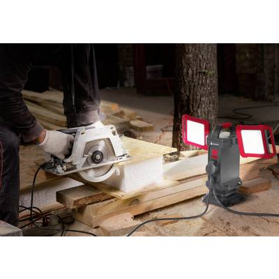 Eine Person sägt Holz mit einer Kreissäge bei Tageslicht, während eine Arbeitslampe mit zwei leuchtenden Leuchten die Szene beleuchtet.