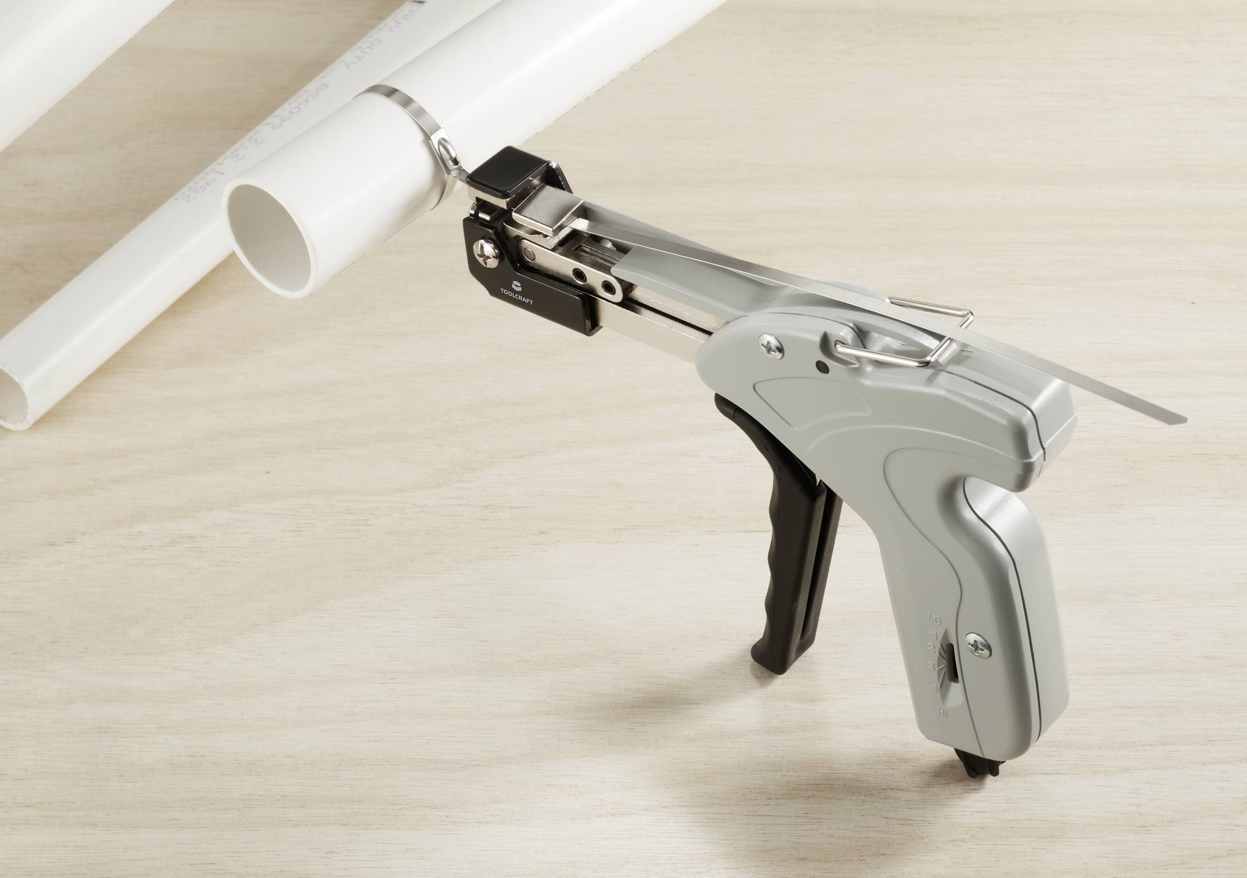 Werkzeug zur Kabelbinderbefestigung neben zwei weißen Kunststoffrohren auf einer Holzoberfläche.