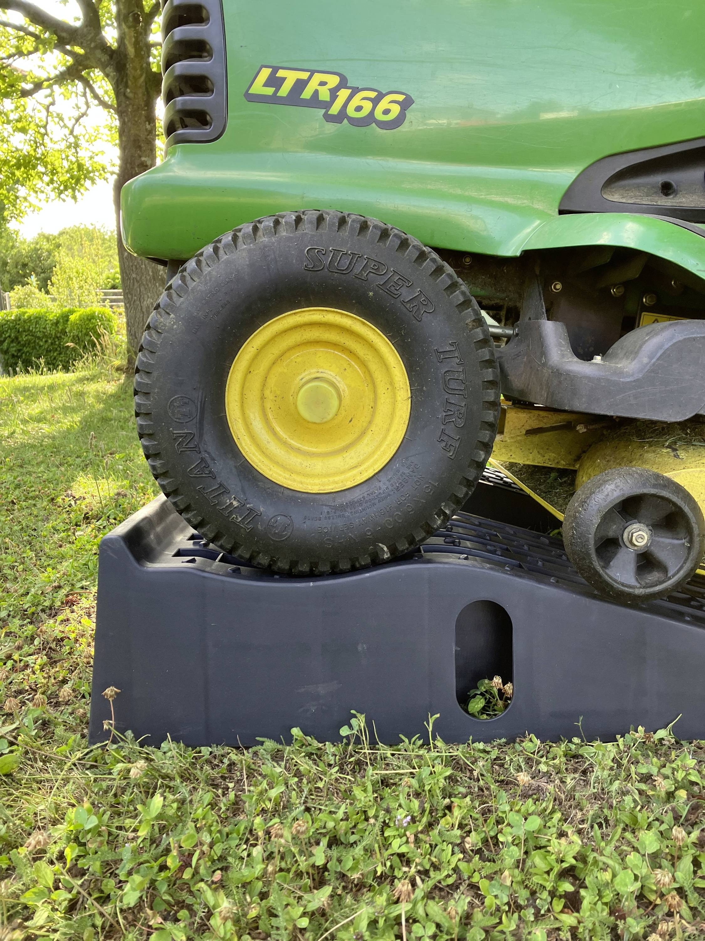 Ein grüner Rasentraktor steht auf einer schwarzen Rampe. Der Vorderreifen mit gelber Felge trägt die Aufschrift 'SUPER TURF'. Im Hintergrund sind Bäume und Gras zu sehen.