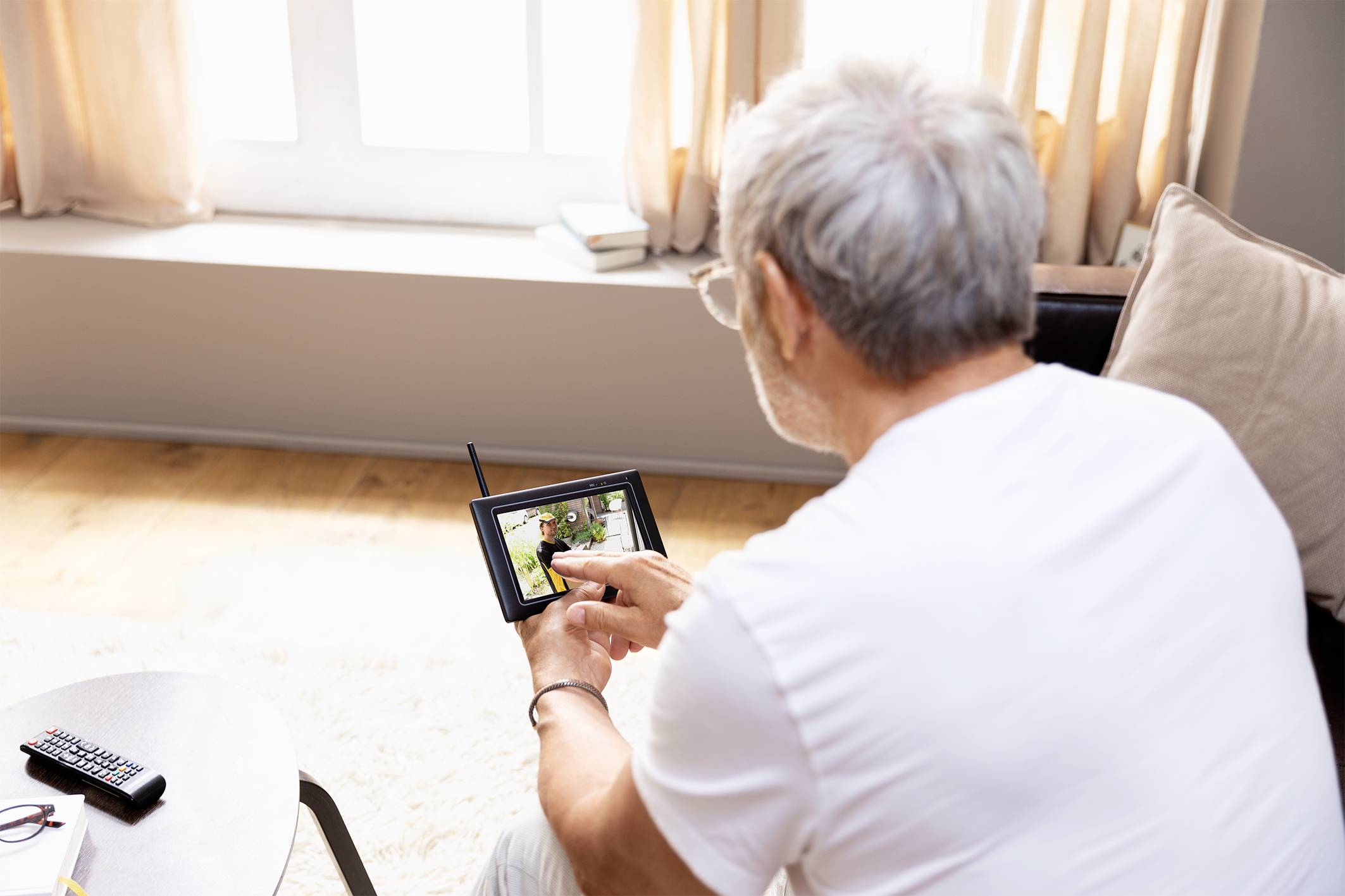 Ein älterer Mann mit grauen Haaren sitzt in einem Wohnzimmer und betrachtet ein Foto auf einem Tablet. Auf dem Tisch liegt eine Fernbedienung.