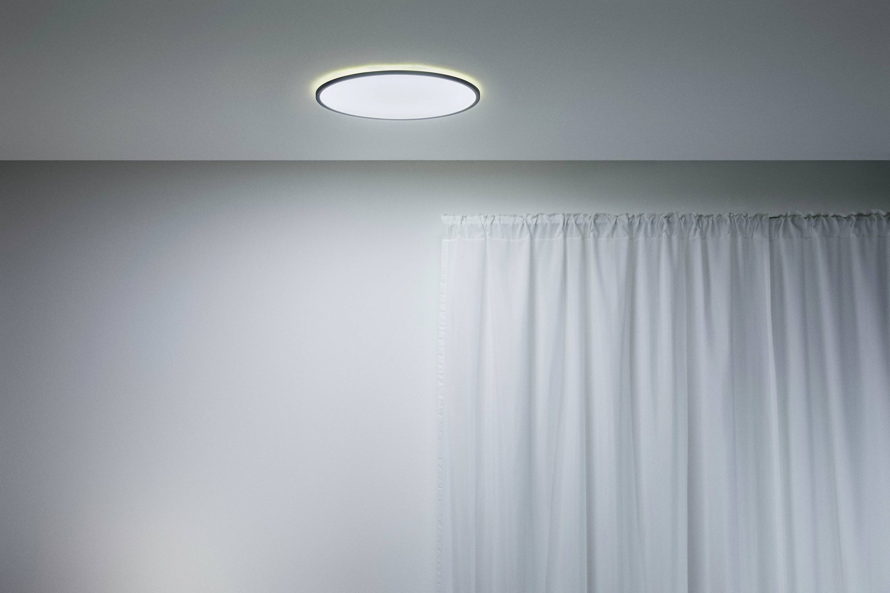 Deckenlampe mit hellem, modernem Design leuchtet einen Raum aus. Rechts ist ein halb geöffneter Vorhang vor einem Fenster zu sehen.