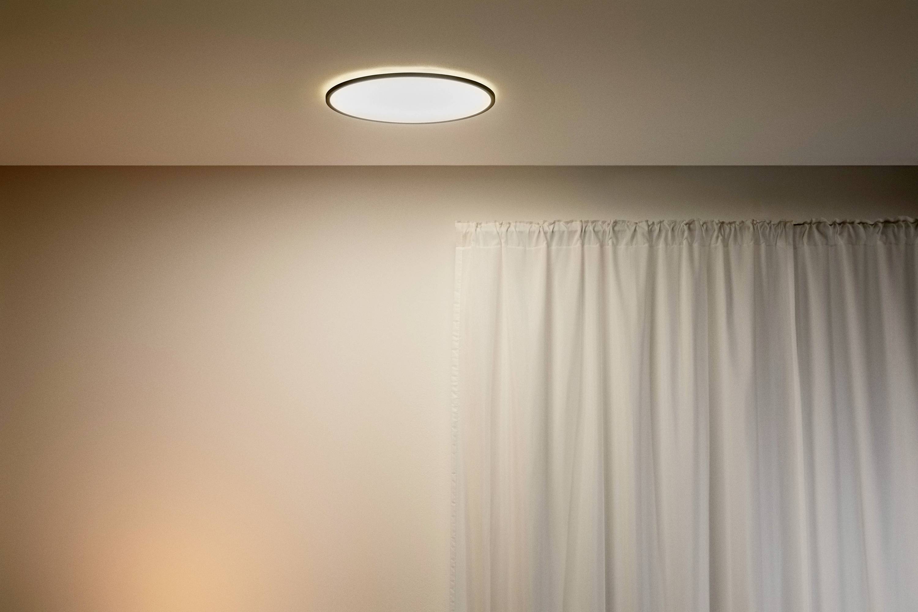 Eine schlichte, runde Deckenleuchte leuchtet sanft neben einem Fenster mit weißen Vorhängen.