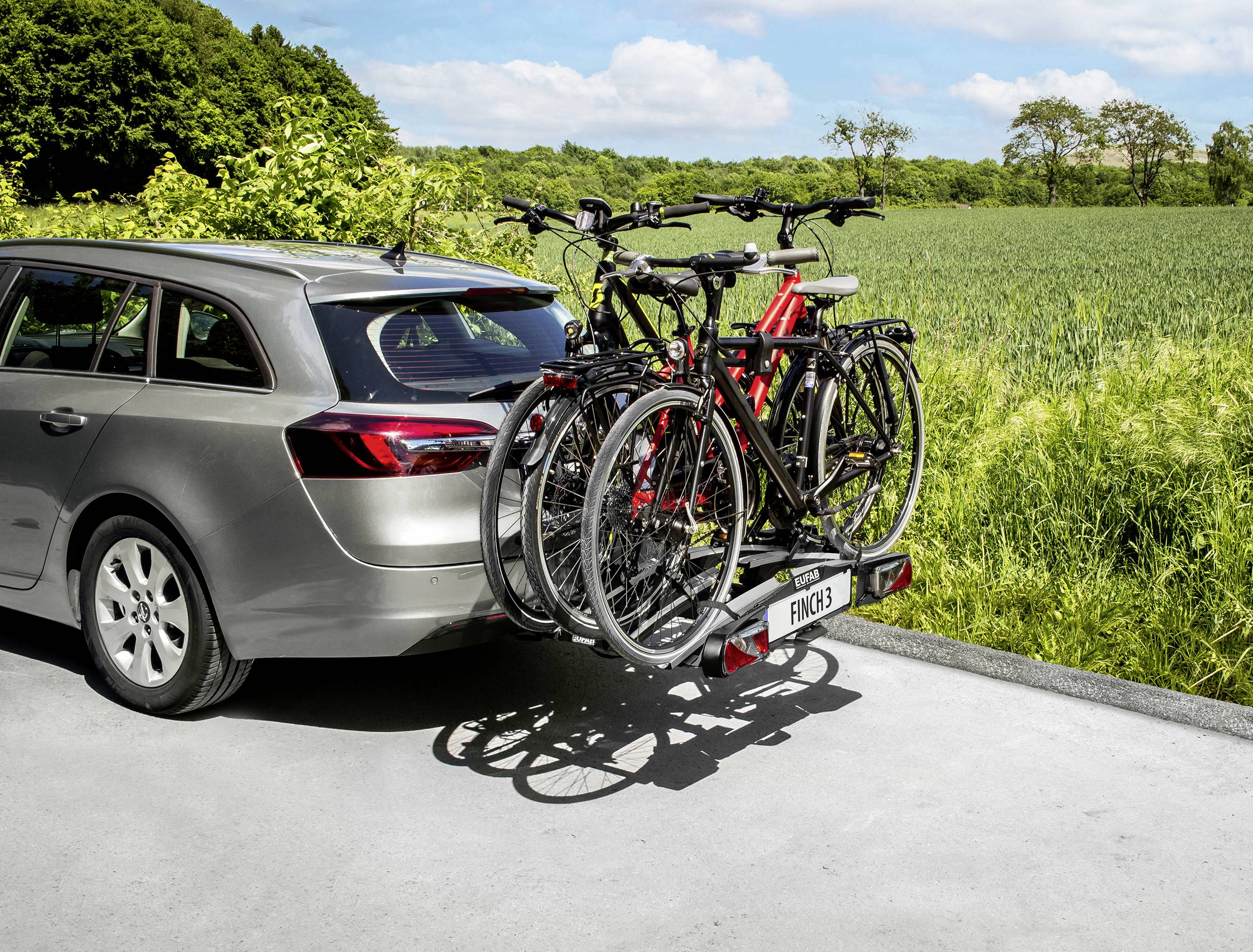 Ein Auto mit drei Fahrrädern auf einem Heckträger steht am Straßenrand neben einem grünen Feld und Bäumen im Hintergrund.