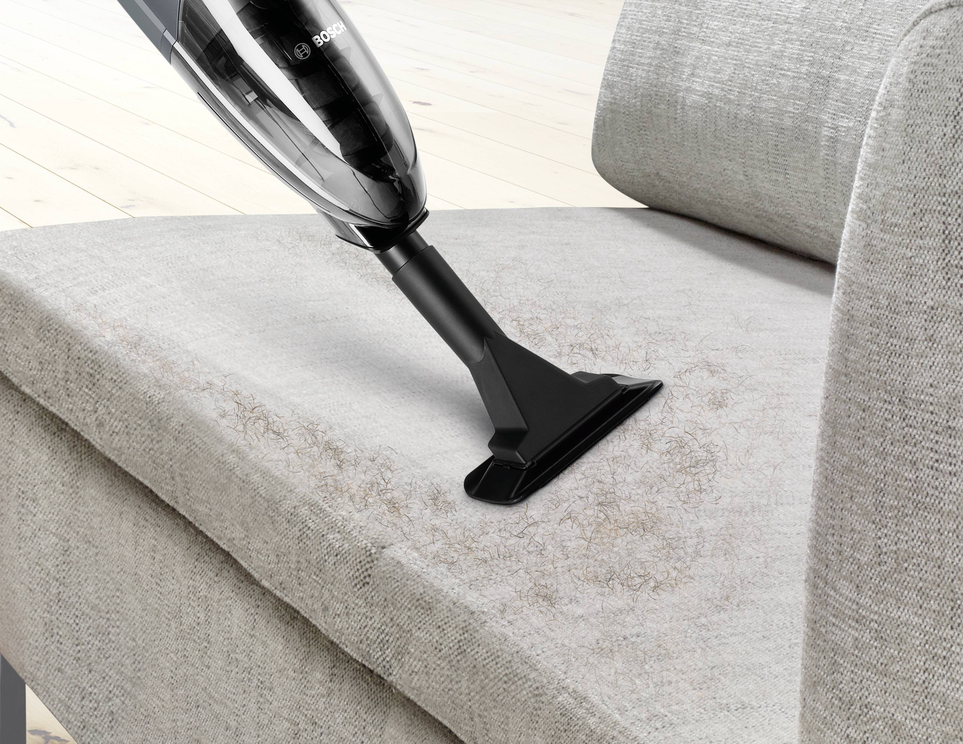 Ein Staubsauger wird verwendet, um Haare und Schmutz von einer grauen Stoffcouch zu entfernen.