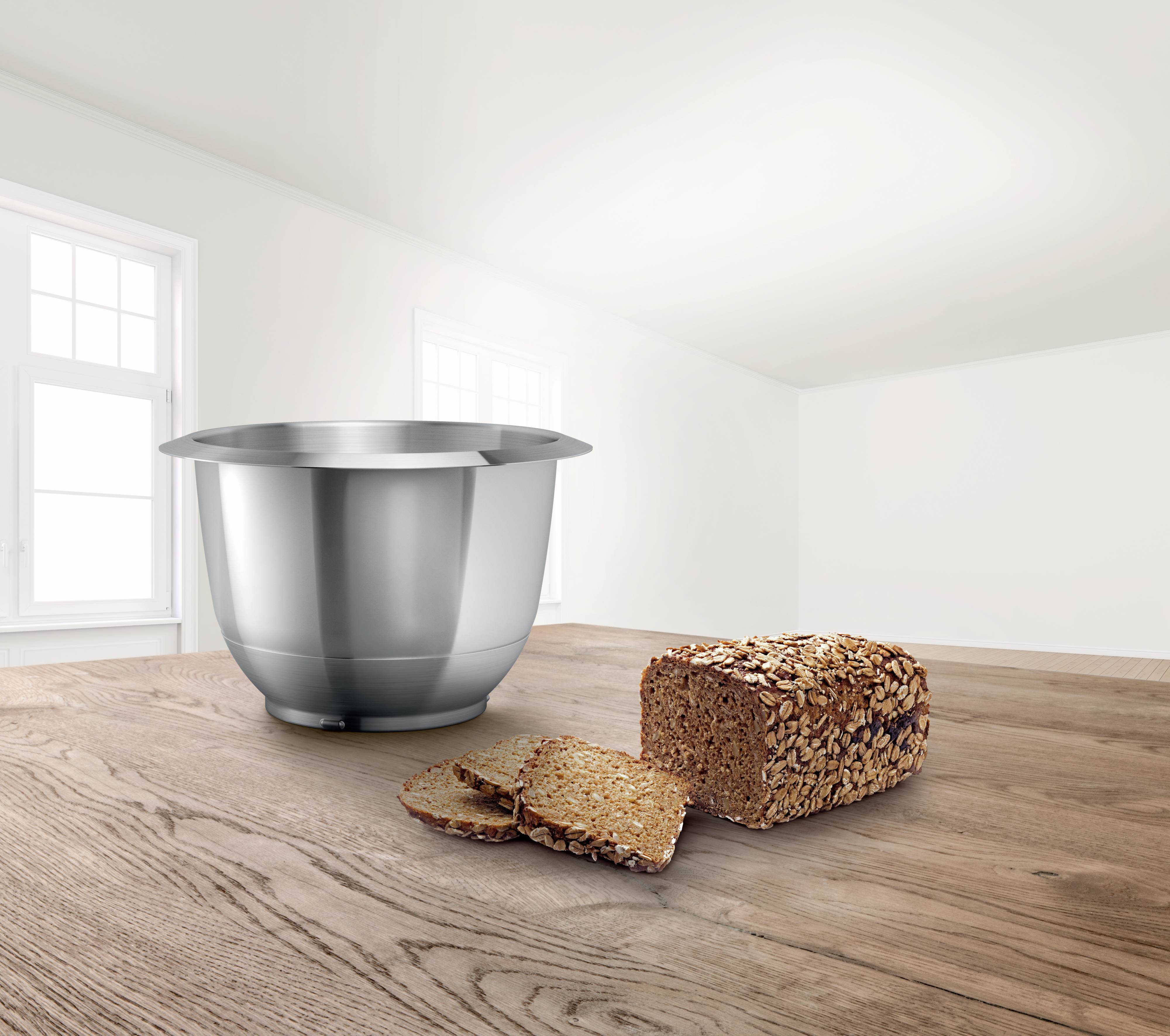 Ein Edelstahlrührschüssel steht auf einem Holztisch neben einem angeschnittenen Laib Vollkornbrot in einem hellen Raum mit großen Fenstern.