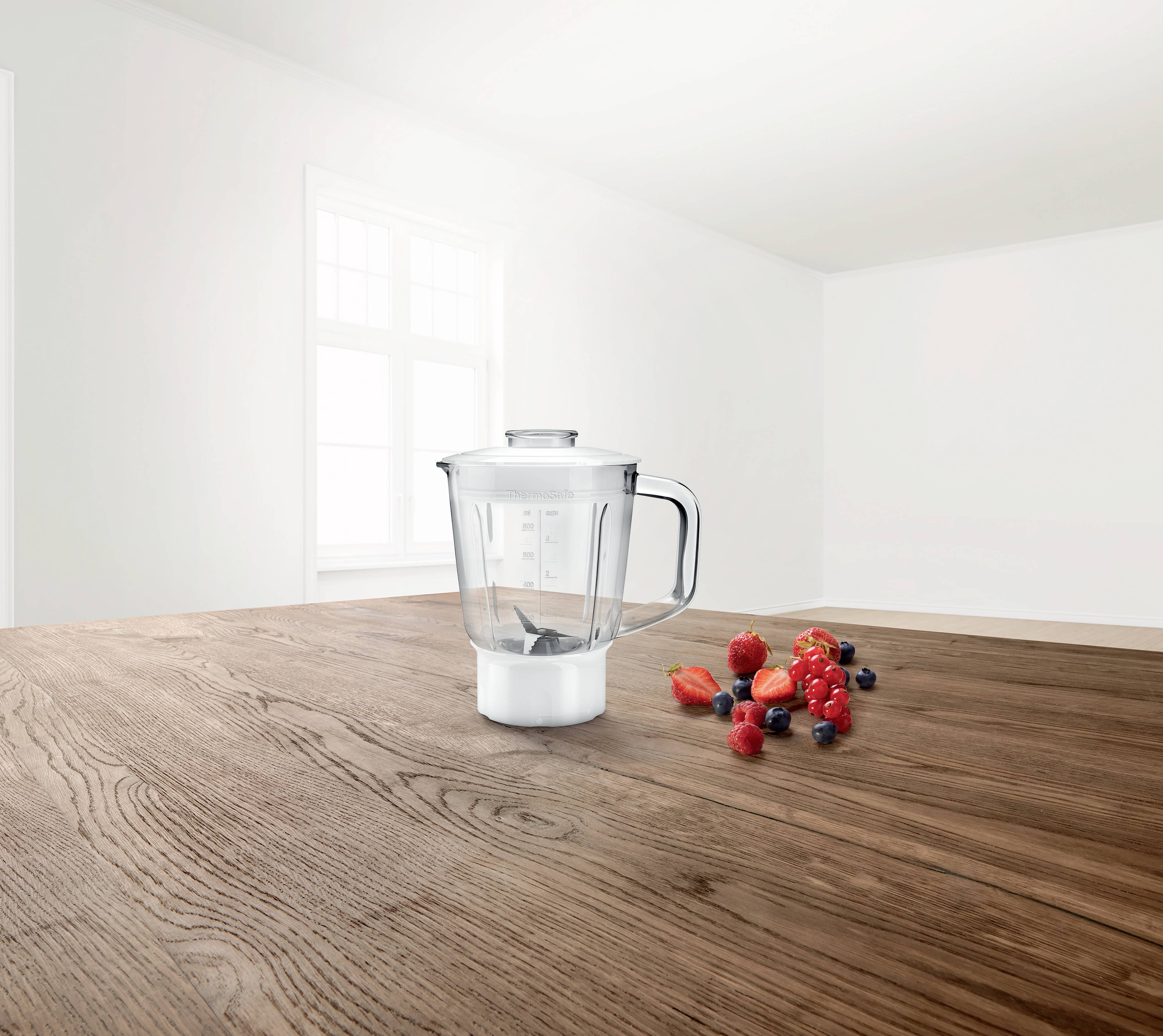 Ein Mixer steht auf einem Holztisch, daneben liegt eine kleine Auswahl an Beerenfrüchten. Im Hintergrund ist ein Fenster.