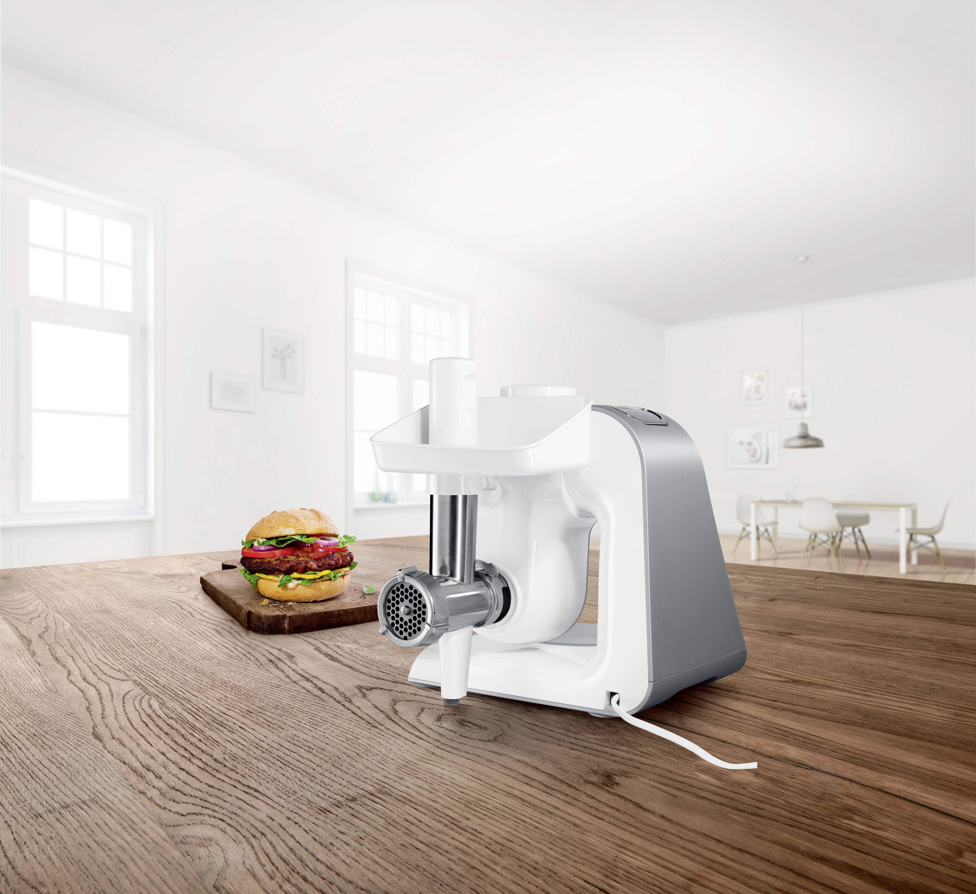 Eine weiße Küchenmaschine steht auf einem hölzernen Tisch in einer modernen, hellen Küche. Daneben liegt ein Burger auf einem Schneidebrett.