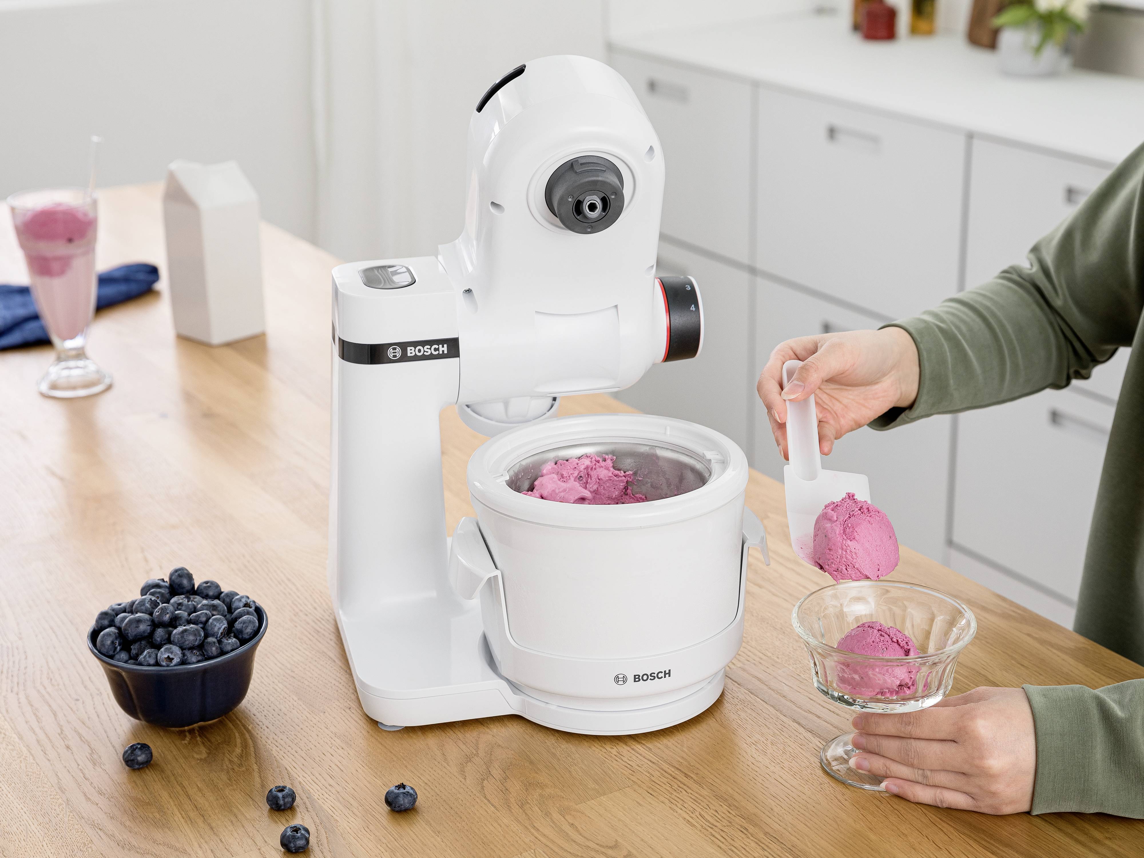 Ein Küchengerät von Bosch mixt rosa Eiscreme. Eine Person serviert das Eis in eine Glasschale. Blaubeeren stehen daneben auf dem Tisch.