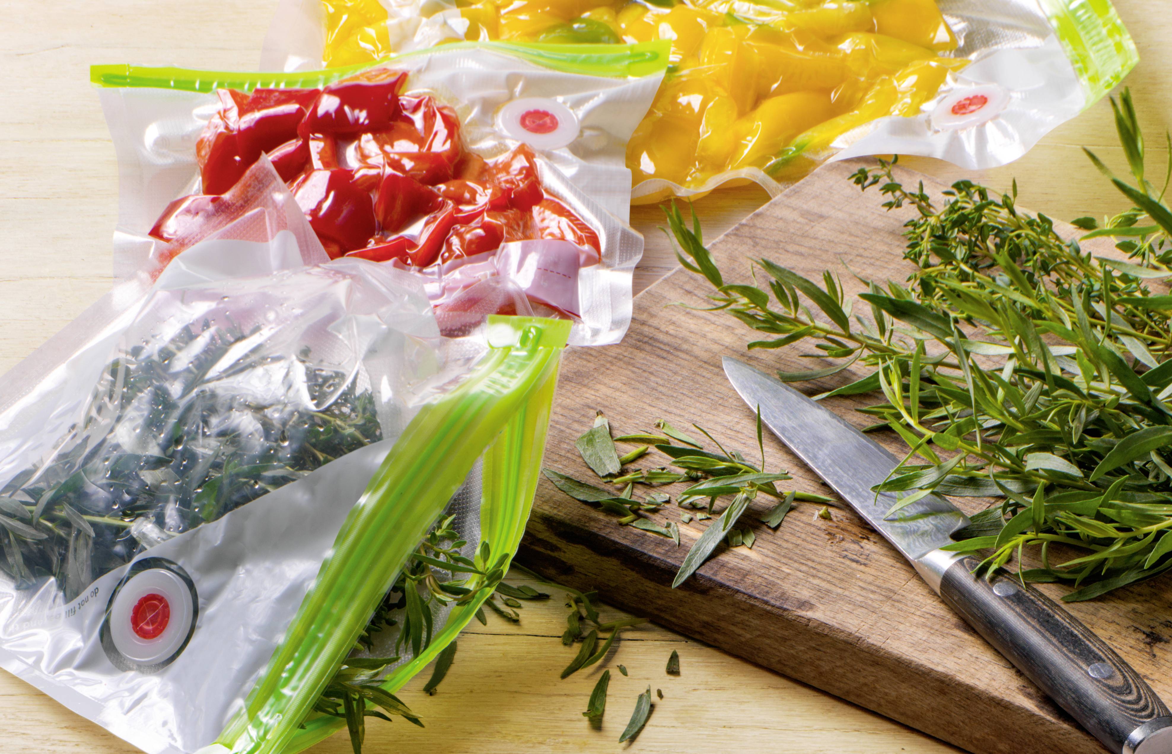 Drei gefüllte Vakuumbeutel mit rotem, gelbem Gemüse und grünen Kräutern liegen auf einem Holzbrett mit Messer und frischen Zweigen.