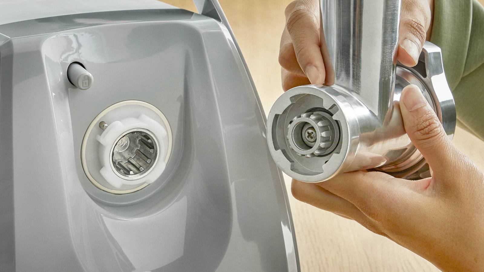 Eine Person montiert ein Metallaufsatz an einen grauen Küchenmixer. Der Aufsatz wird in die vorgesehene Öffnung eingesetzt.