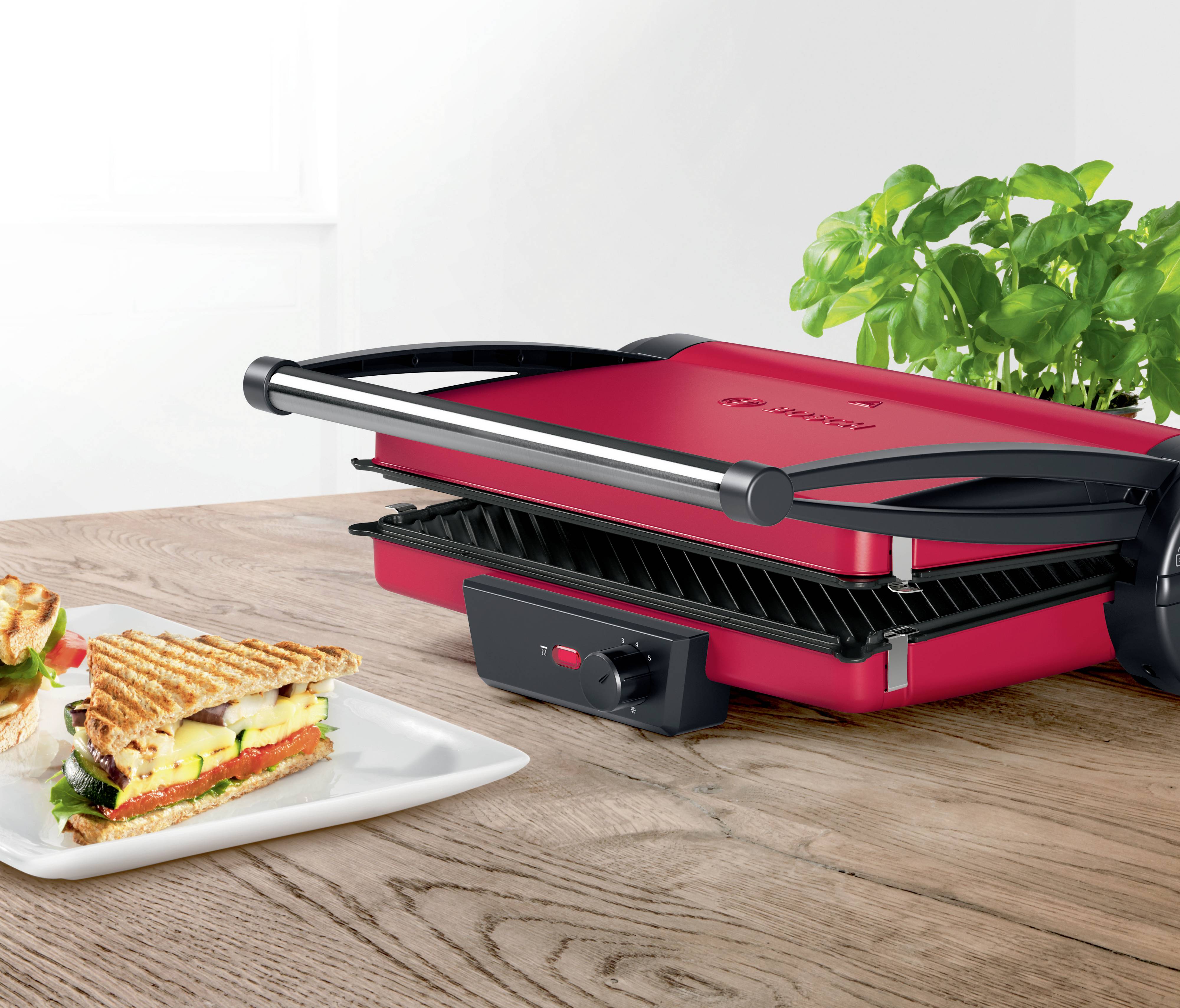 Ein roter Kontaktgrill auf einem Holztisch neben frischem Basilikum, mit einem gegrillten Sandwich auf einem Teller davor.