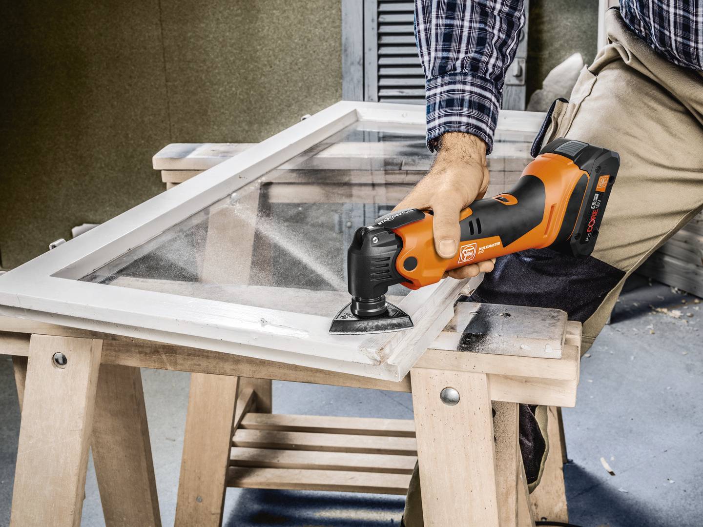 Eine Person schleift mit einem orange-schwarzen Multitool das Holz eines auf Böcken liegenden Fensterrahmens ab.