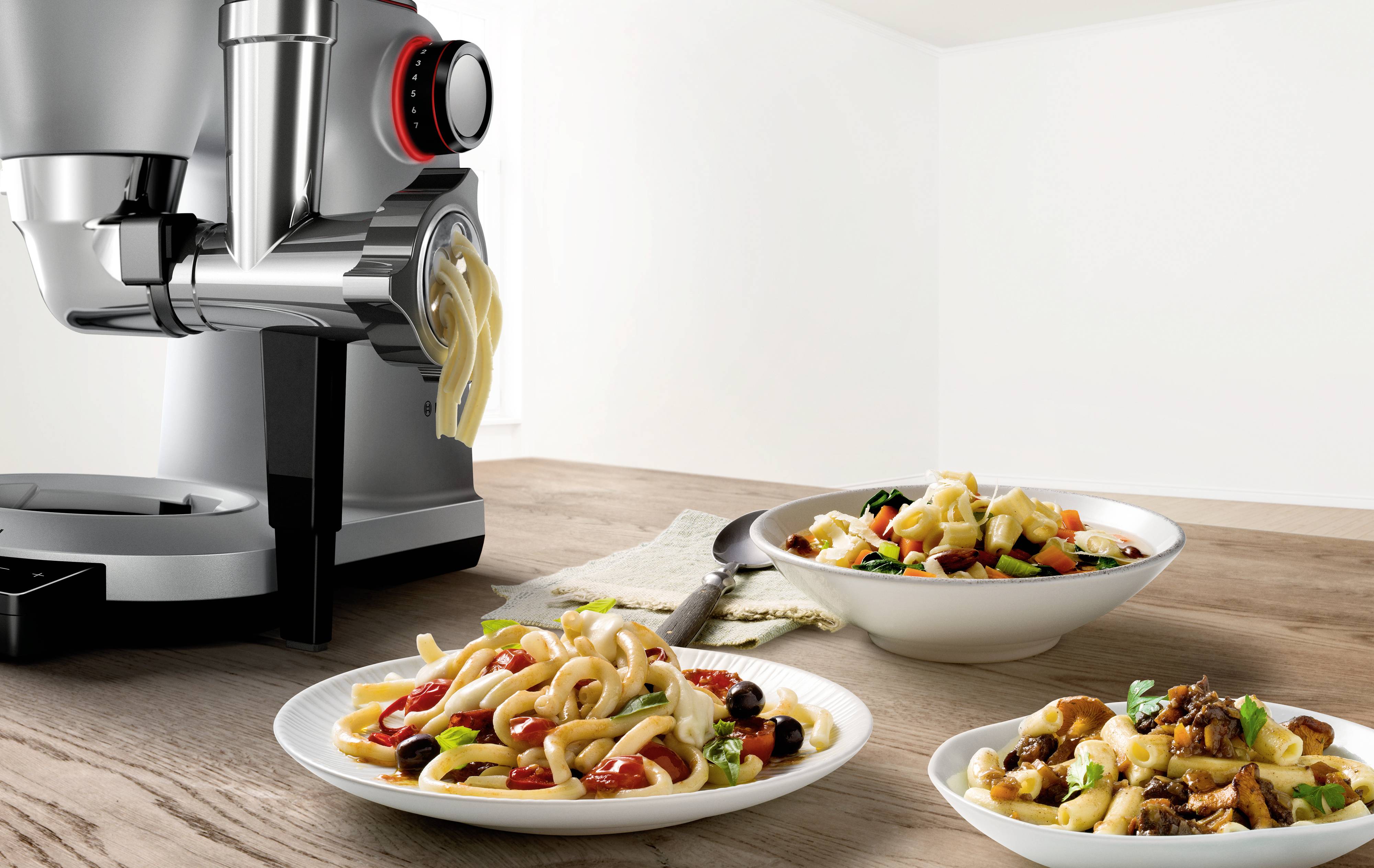 Nudelmaschine presst frische Pasta. Drei Teller mit unterschiedlichen Nudelgerichten auf einem Holztisch.