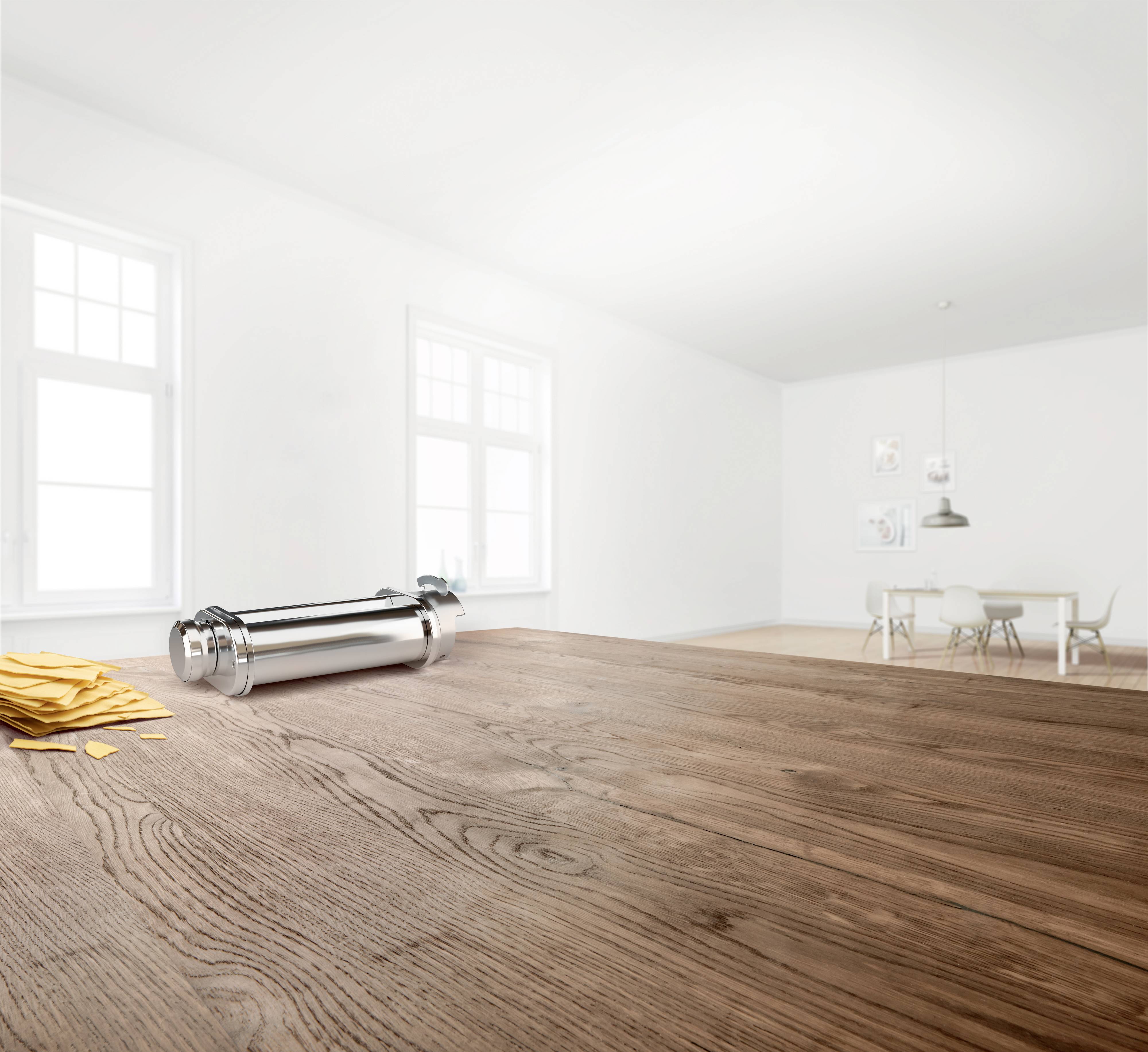 Schlichter, heller Raum mit Holztisch im Vordergrund. Auf dem Tisch liegen gelbe Papierbögen neben einer silbernen Thermoskanne. Im Hintergrund ein Esstisch mit Stühlen.