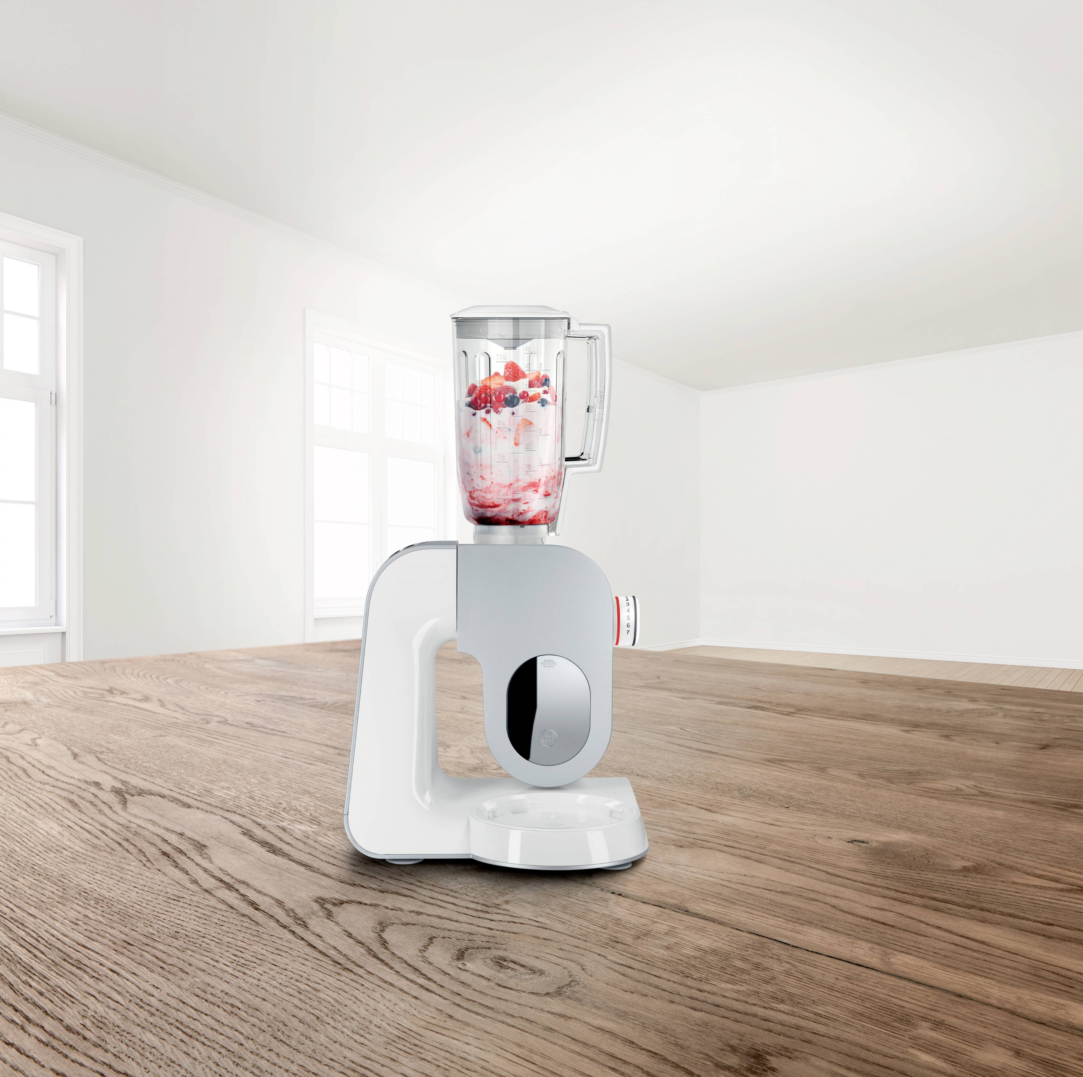 Ein weißer Mixer steht auf einem Holztisch in einem hellen Raum. Im Behälter sind rote und weiße Zutaten für einen Smoothie zu sehen.