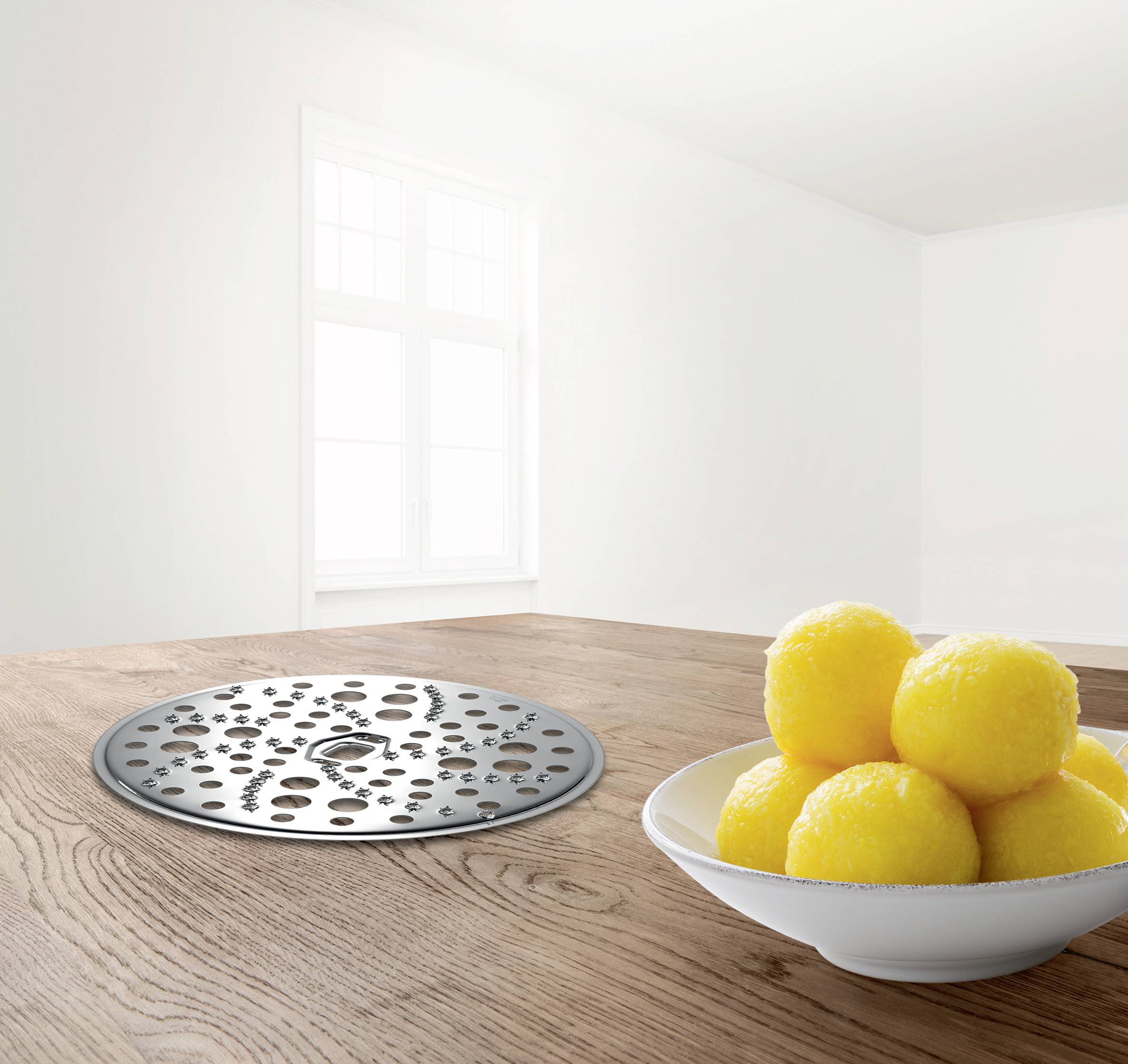 Ein weißer Raum mit einem Holztisch, darauf eine Metallscheibe mit Löchern und eine Schale mit gelben Bällen. Helle Fenster im Hintergrund.