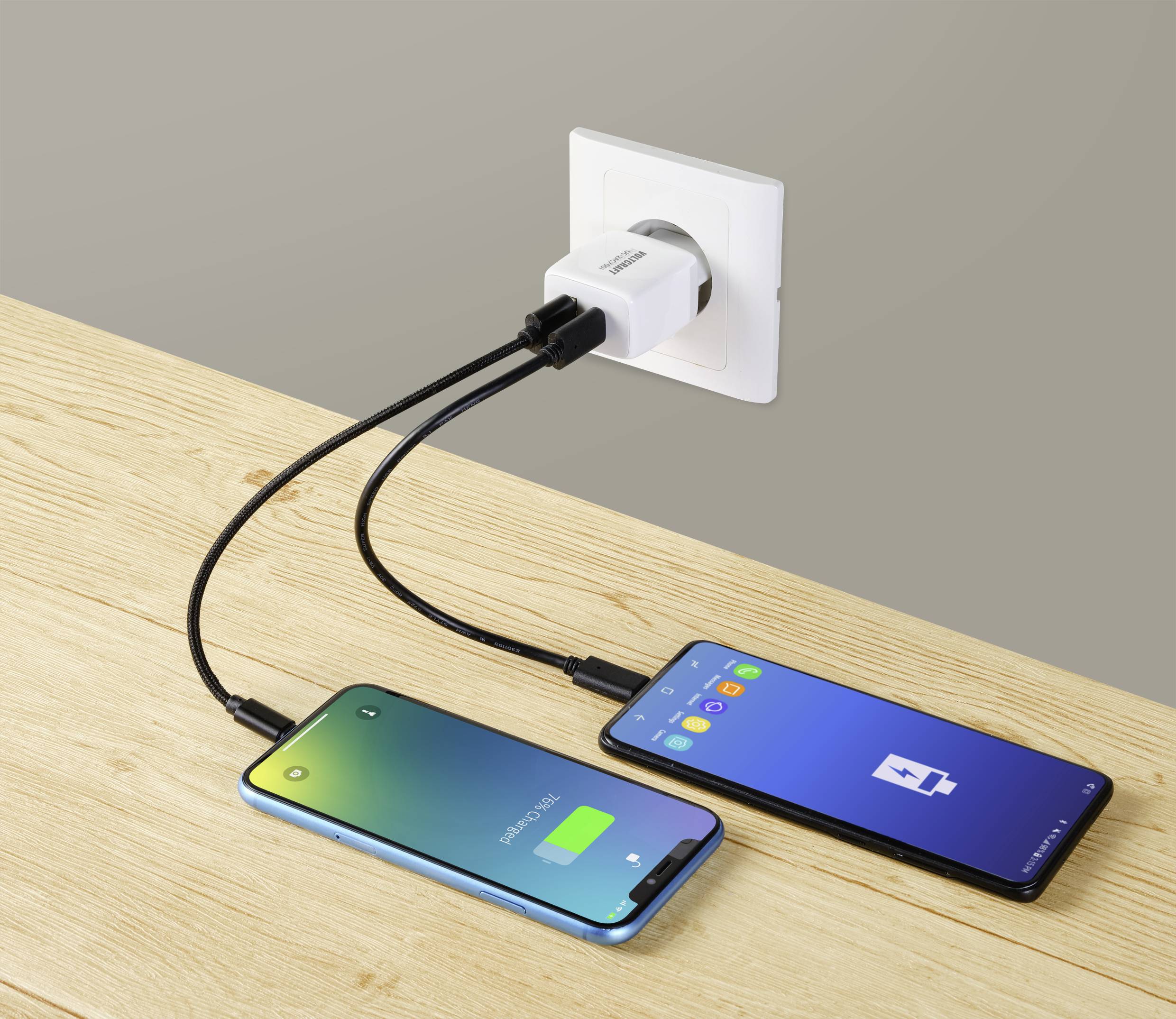 Zwei Handys werden auf einem Tisch über ein duales USB-Ladegerät in einer Steckdose aufgeladen; eines ist fast voll, das andere niedrig.