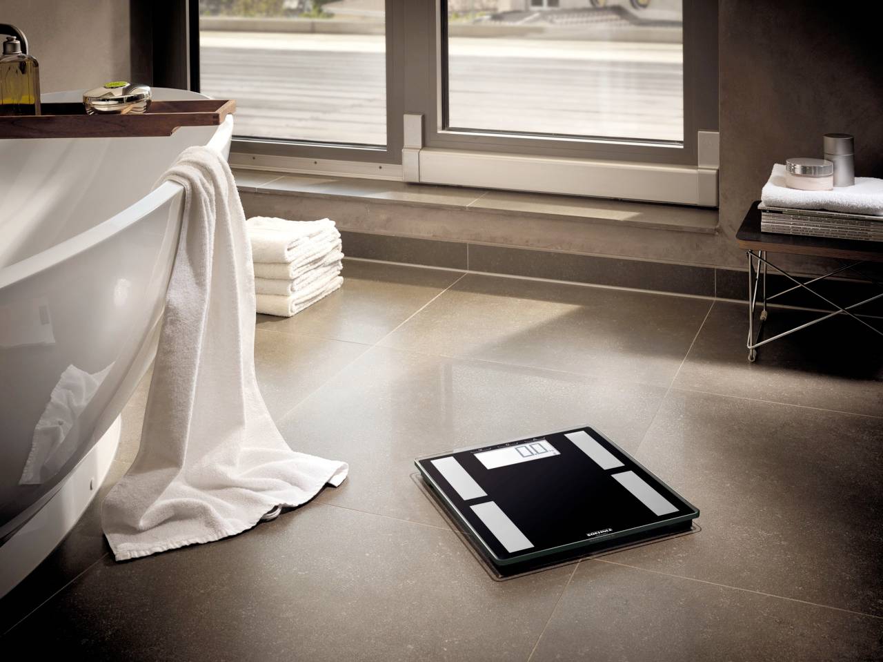 Eine digitale Personenwaage auf einem Badezimmerboden, neben der Badewanne. Durch das Fenster fällt Tageslicht in den Raum.