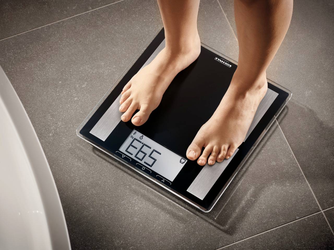 Person steht auf einer digitalen Waage, die 66,5 Kilogramm anzeigt. Die Waage liegt auf einem gefliesten Boden.
