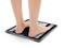 Eine Person steht barfuß auf einer digitalen Waage. Das Display zeigt das Gewicht in Kilogramm an.
