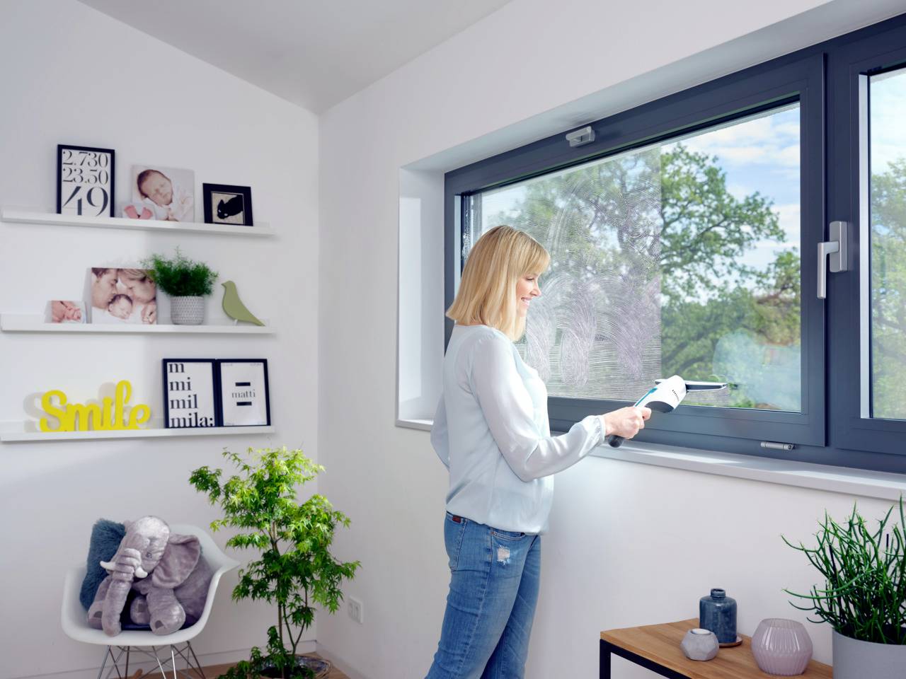 Eine Frau wischt ein Fenster in einem hellen Raum. Im Hintergrund sind Pflanzen, Bilderrahmen und Regale mit Dekorationen zu sehen.
