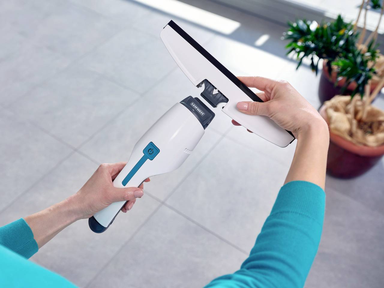 Eine Person hält einen elektrischen Fensterreiniger in der Hand, bereit, eine Glasoberfläche zu reinigen. Im Hintergrund ist eine Pflanze sichtbar.