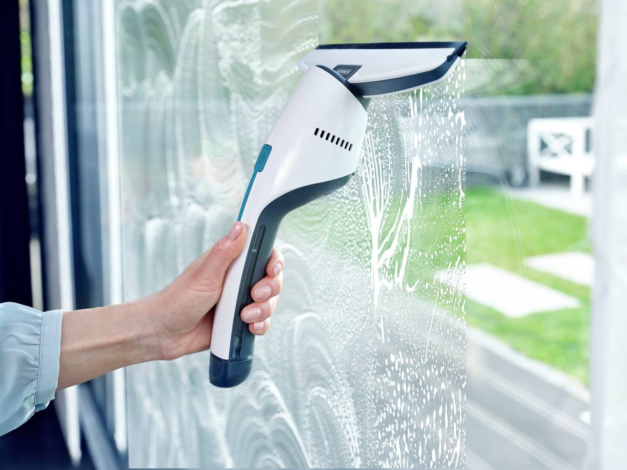 Eine Hand hält einen Fenstersauger, der Seifenwasser von einer Glasscheibe entfernt, im Hintergrund ist ein Garten zu sehen.
