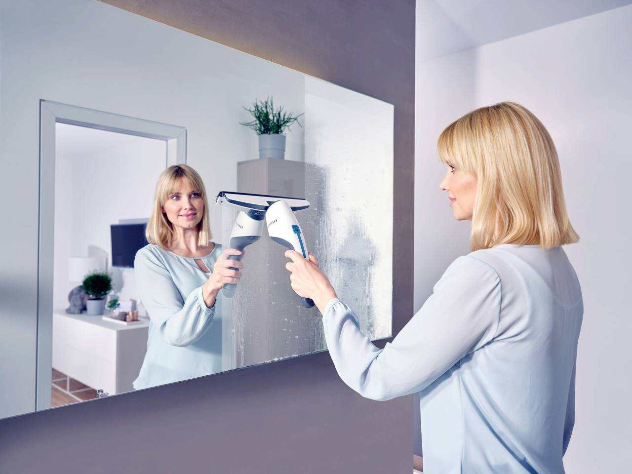 Frau reinigt einen Spiegel mit einem Fensterabzieher; modernes Interieur mit Pflanzen und TV im Hintergrund reflektiert.