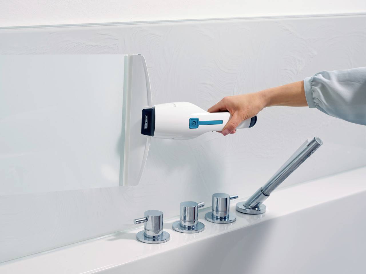Eine Person hält einen modernen, weißen Handstaubsauger über einer weißen Fläche neben einem Wasserhahn mit drei Drehknöpfen.