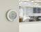 Ein digitaler Thermostat an einer Wand zeigt eine Temperatur von 24,5°C und eine Luftfeuchtigkeit von 56% an, im Hintergrund ein Büro.