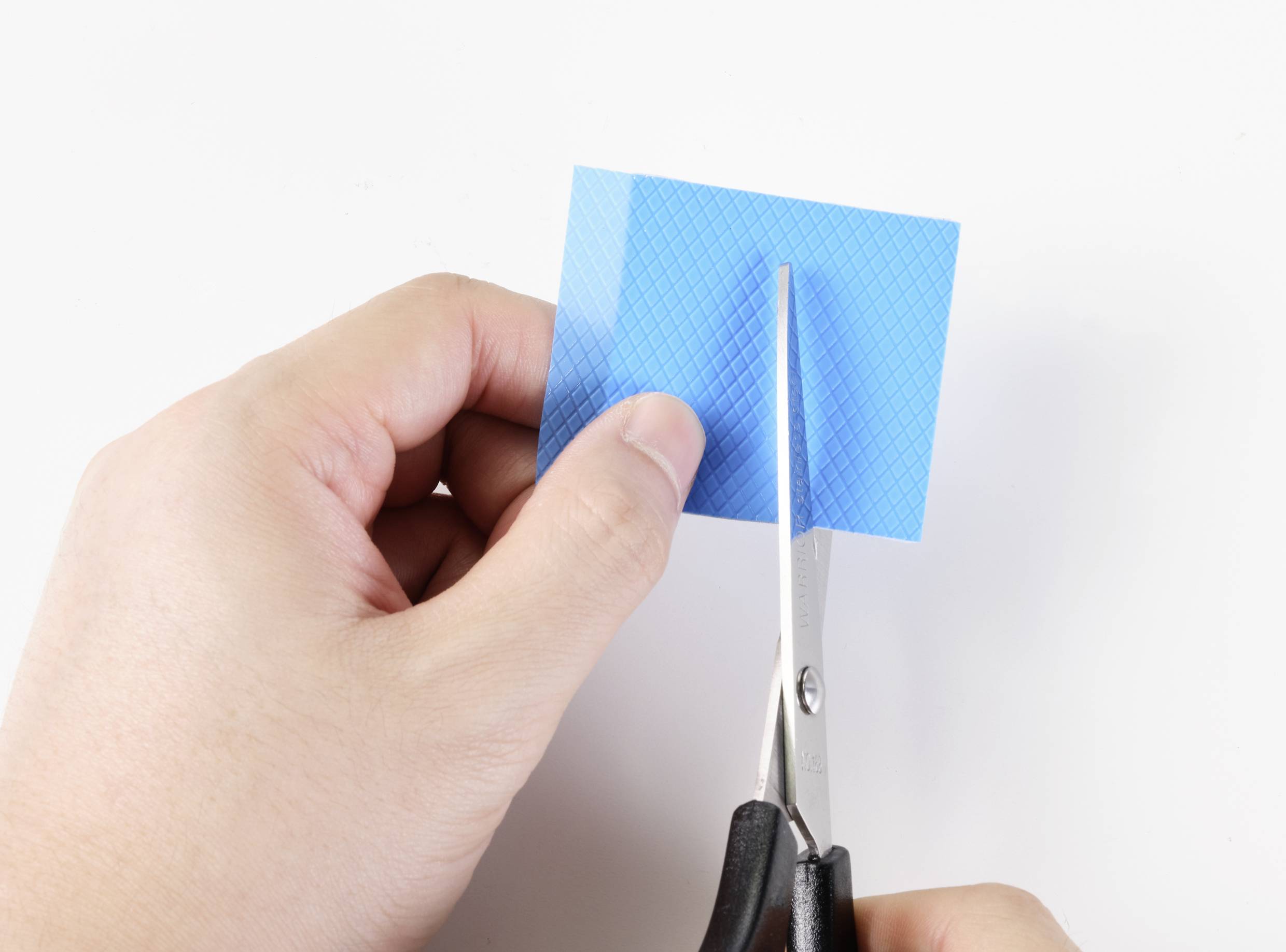 Eine Hand benutzt eine Schere, um ein quadratisches Stück blauen Karton zu schneiden.