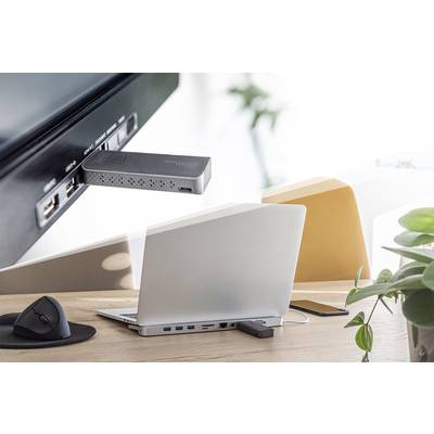 Ein Laptop steht auf einem Schreibtisch mit einer Dockingstation und Zubehör. Ein Fernseher zeigt eine verbundene Streaming-Box. Pflanzen im Hintergrund.