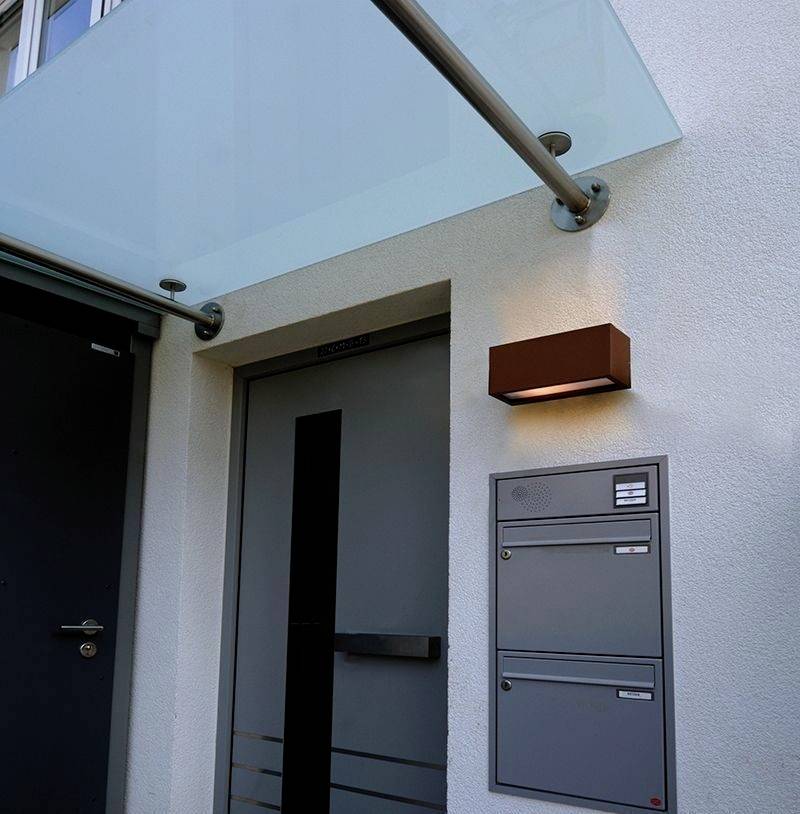 Eingang eines modernen Gebäudes mit einer Glastür, einem Briefkasten und einer Außenleuchte auf der rechten Seite.
