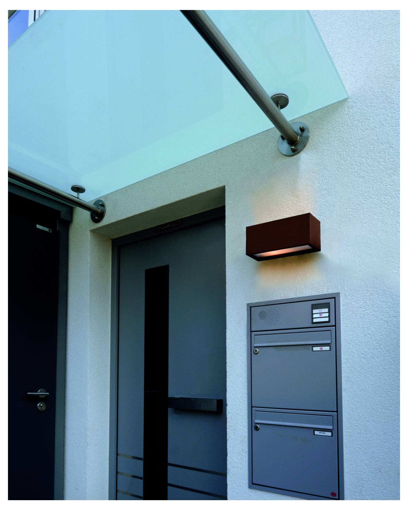 Hauseingang mit blauem Vordach, zwei Türen, Wandleuchte und einer Reihe von Briefkästen an der Außenwand.
