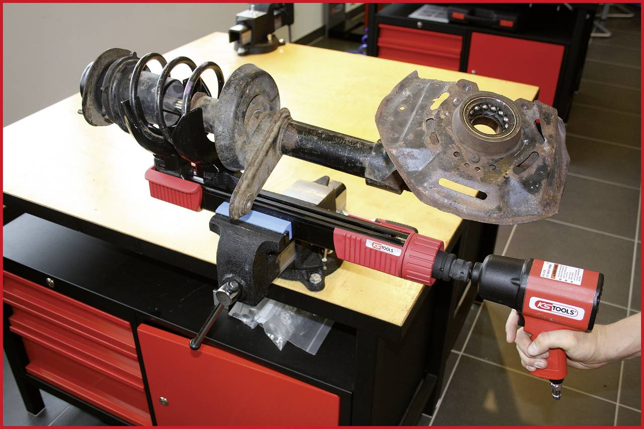 Ein Stoßdämpfer wird mit einem Schlagbohrer auf einer Werkbank bearbeitet. Im Hintergrund sind rote Schubladen zu sehen.