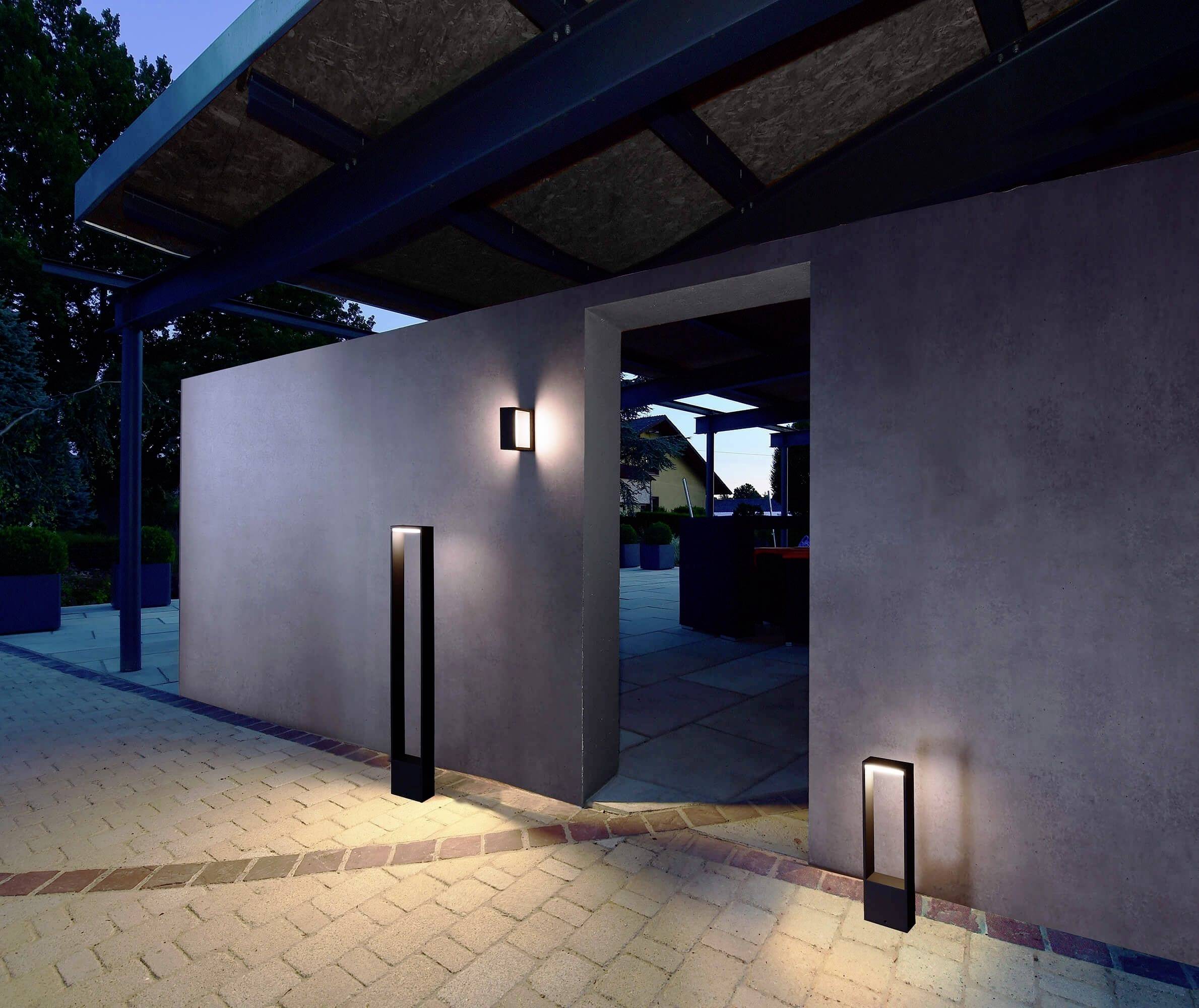 Ein moderner Eingang abends mit stilvoller Beleuchtung. Dunkle Wandleuchten und Bodenlampen betonen den minimalistischen Betonaufbau.