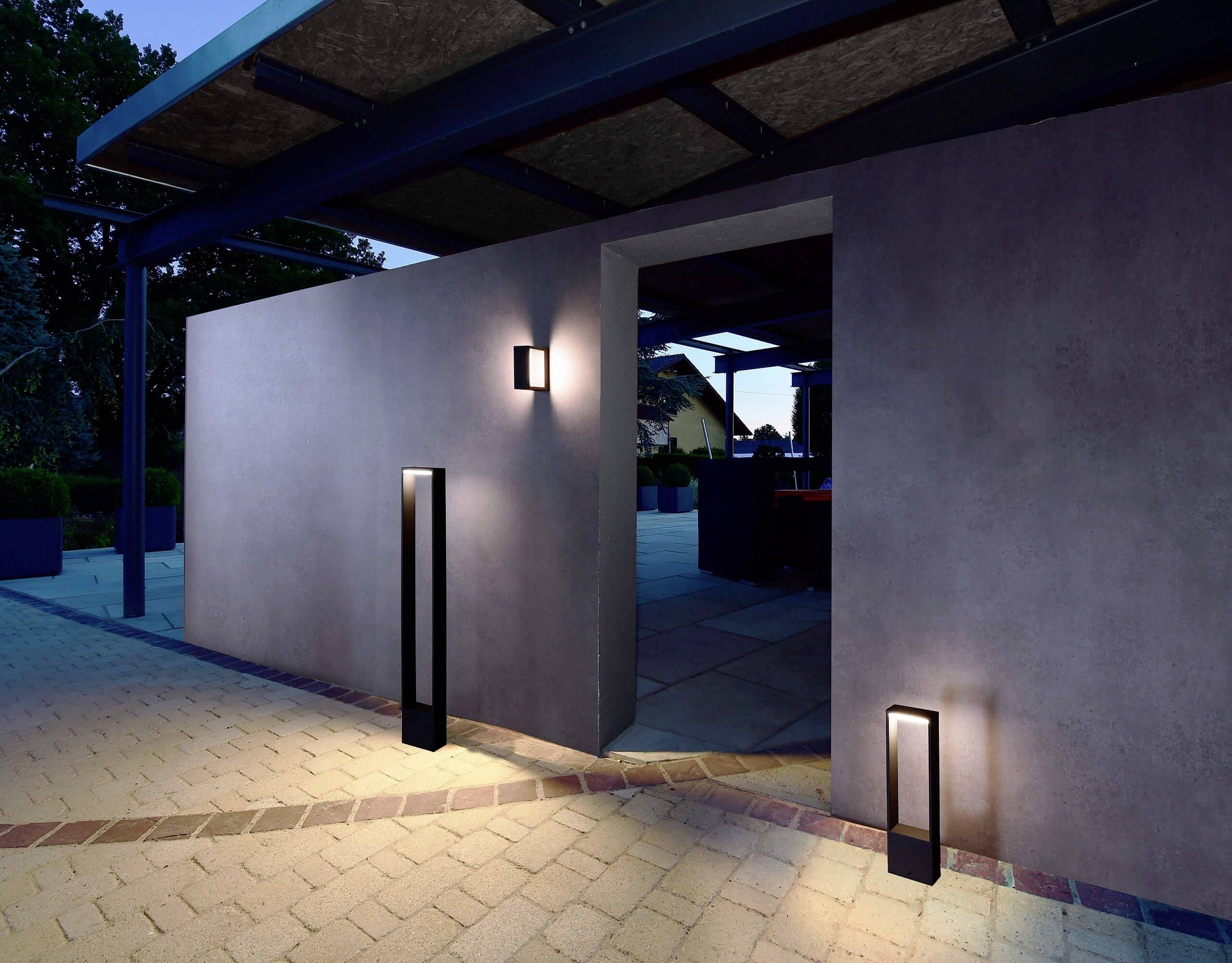 Eine moderne Außenbeleuchtung sorgt für stimmungsvolle Akzente an einer grauen Betonwand und einem gepflasterten Weg bei Dämmerung.