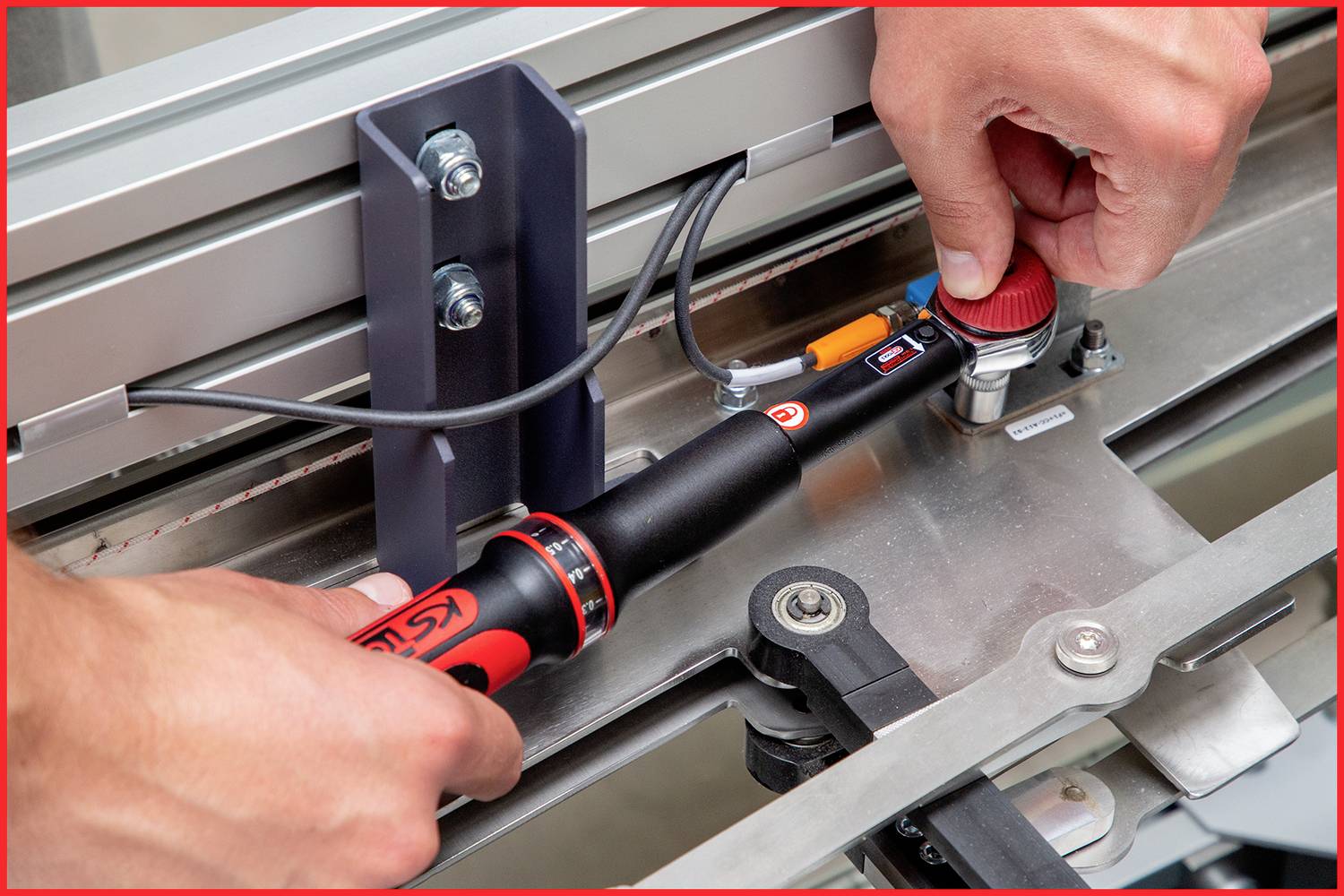 Zwei Hände bedienen ein Drehmomentwerkzeug an einer Metallvorrichtung mit Kabeln und Schraubbefestigungen, Nahaufnahme.