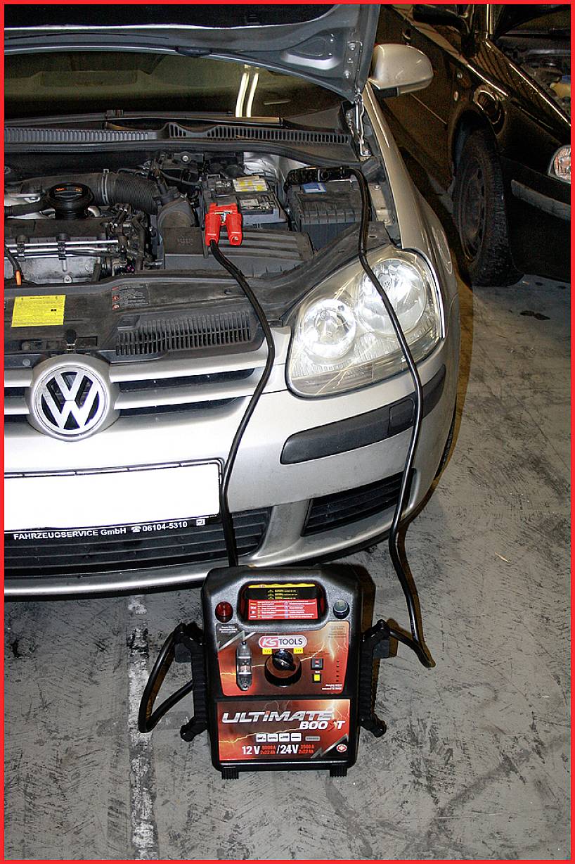 Auto mit geöffneter Motorhaube, angeschlossen an ein tragbares Starthilfegerät in einer Garage.