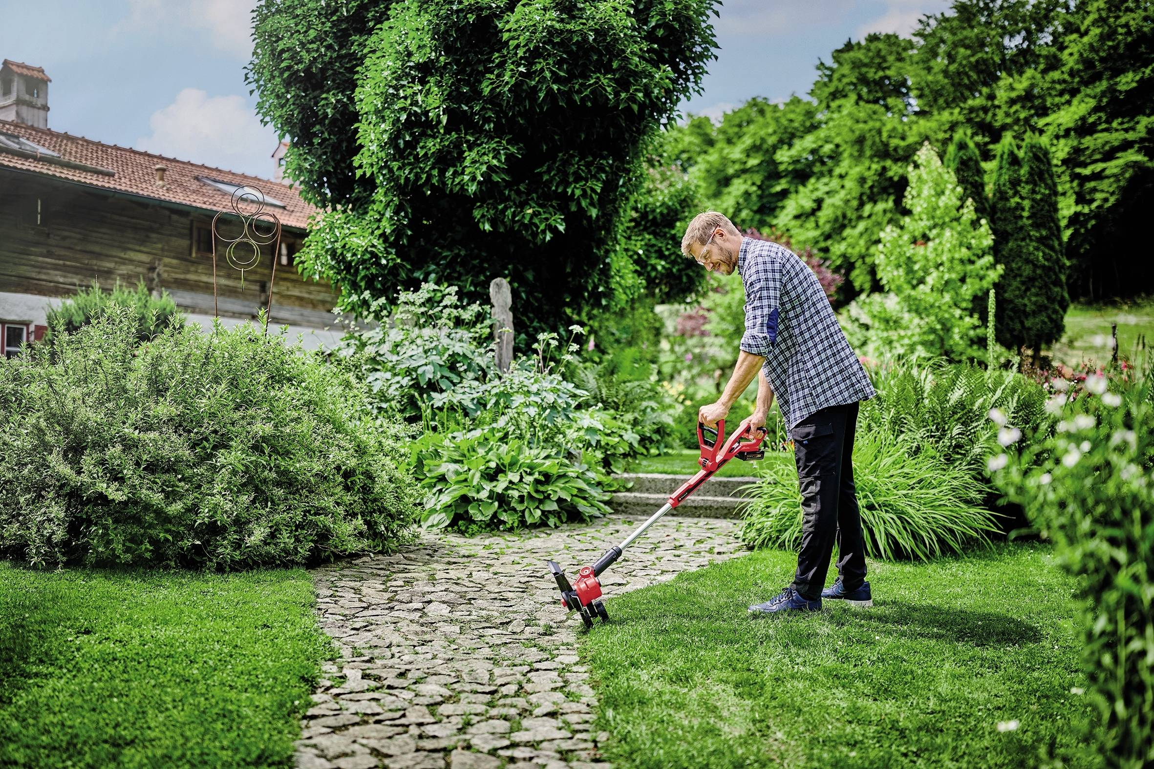 Ein Mann mäht den Rasen mit einem elektrischen Trimmer auf einem gepflegten Gartenweg, umgeben von grünem Laub und einem Holzhaus im Hintergrund.